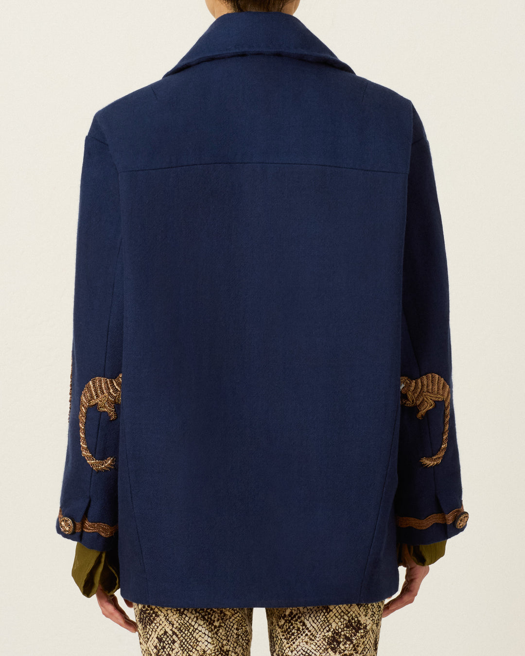 Wearing the Rivoli Midnight Mischief Coat, featuring brown embroidered lizard designs on the sleeves, shown from the back and paired with snake print pants.