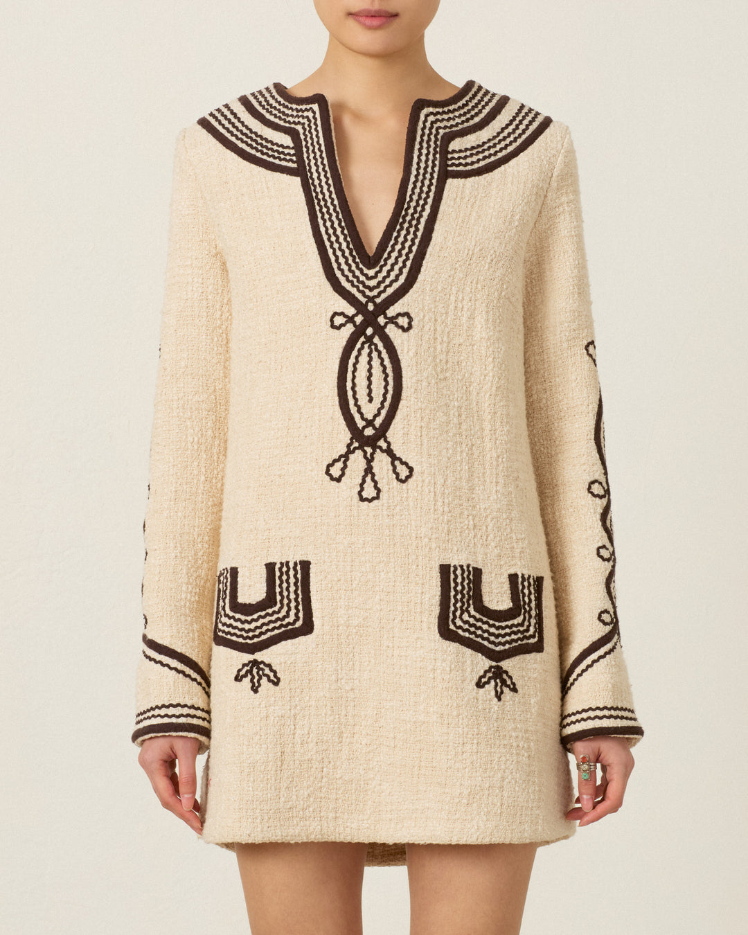 A woman wearing the Rosie Bushland Cord Dress—a cream-colored, hand-embroidered, long-sleeve dress with black patterns on the neckline, cuffs, pockets, and sleeves—stands against a neutral background.