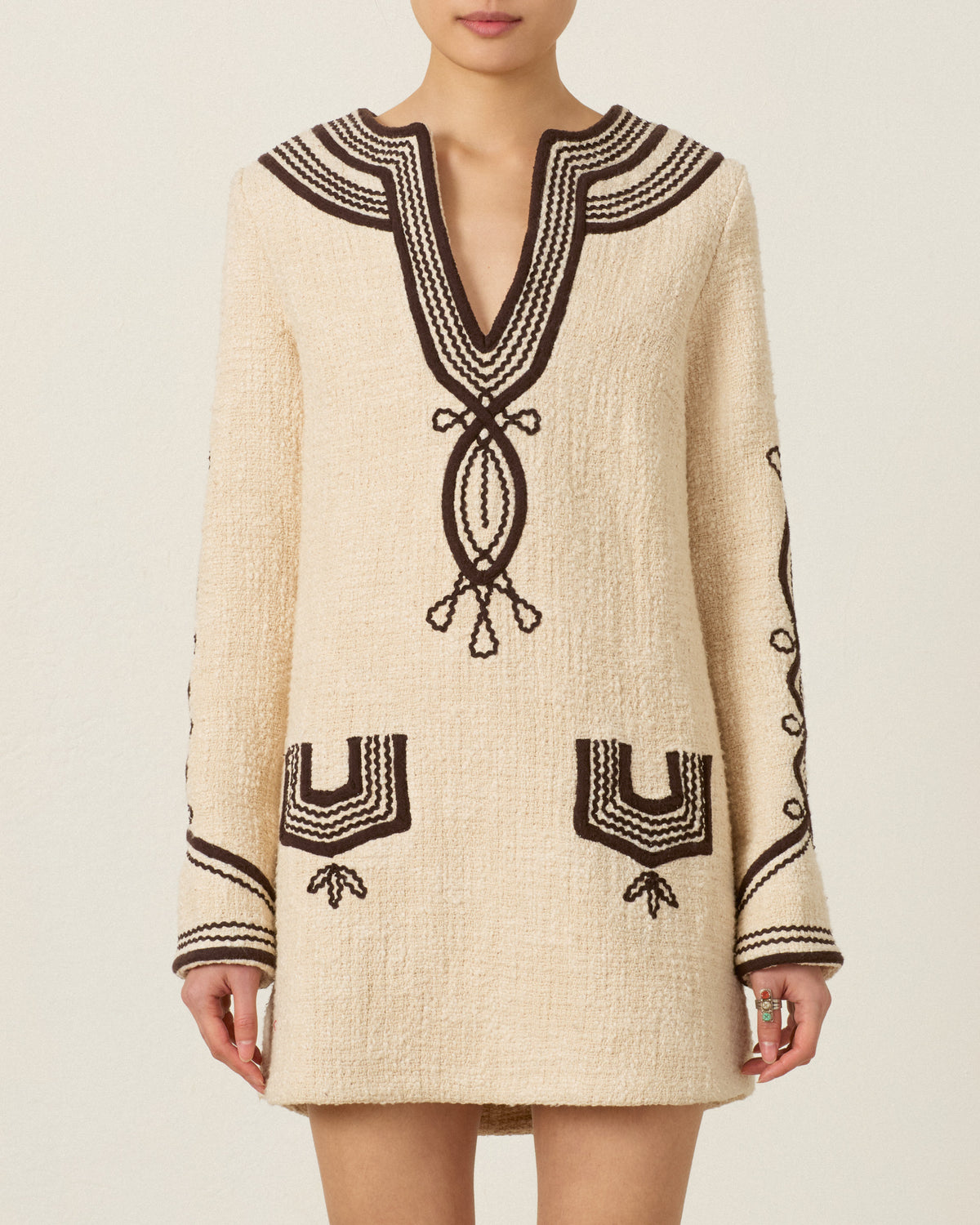 A woman wearing the Rosie Bushland Cord Dress—a cream-colored, hand-embroidered, long-sleeve dress with black patterns on the neckline, cuffs, pockets, and sleeves—stands against a neutral background.
