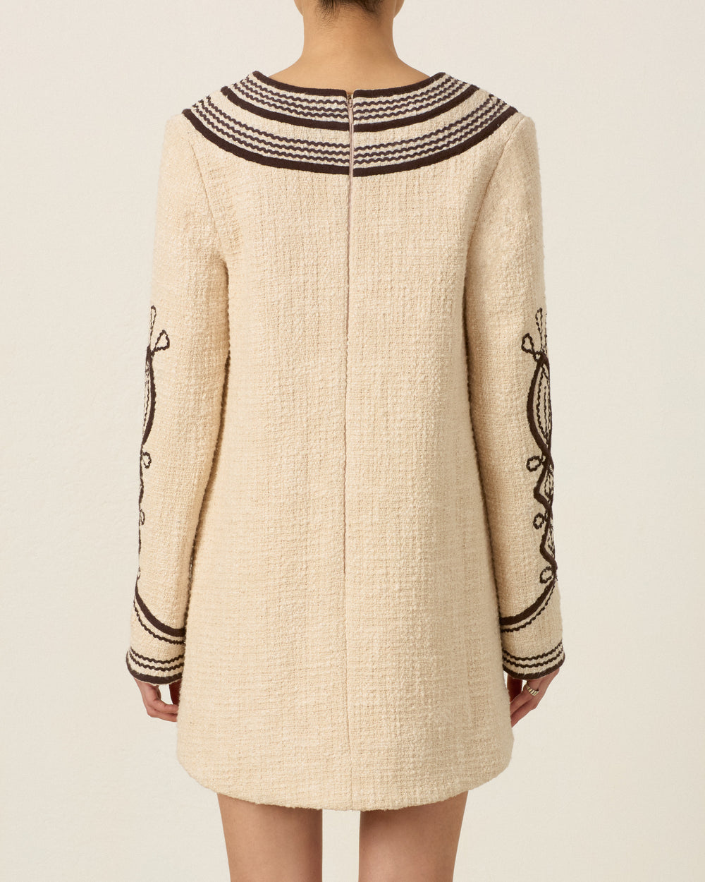 Model wearing the Rosie Bushland Cord Dress, a cream-colored, hand-embroidered long-sleeve dress with dark brown soutache embroidery trim and detailing, shown from the back.