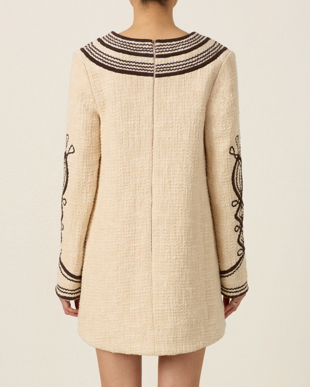 Model wearing the Rosie Bushland Cord Dress, a cream-colored, hand-embroidered long-sleeve dress with dark brown soutache embroidery trim and detailing, shown from the back.