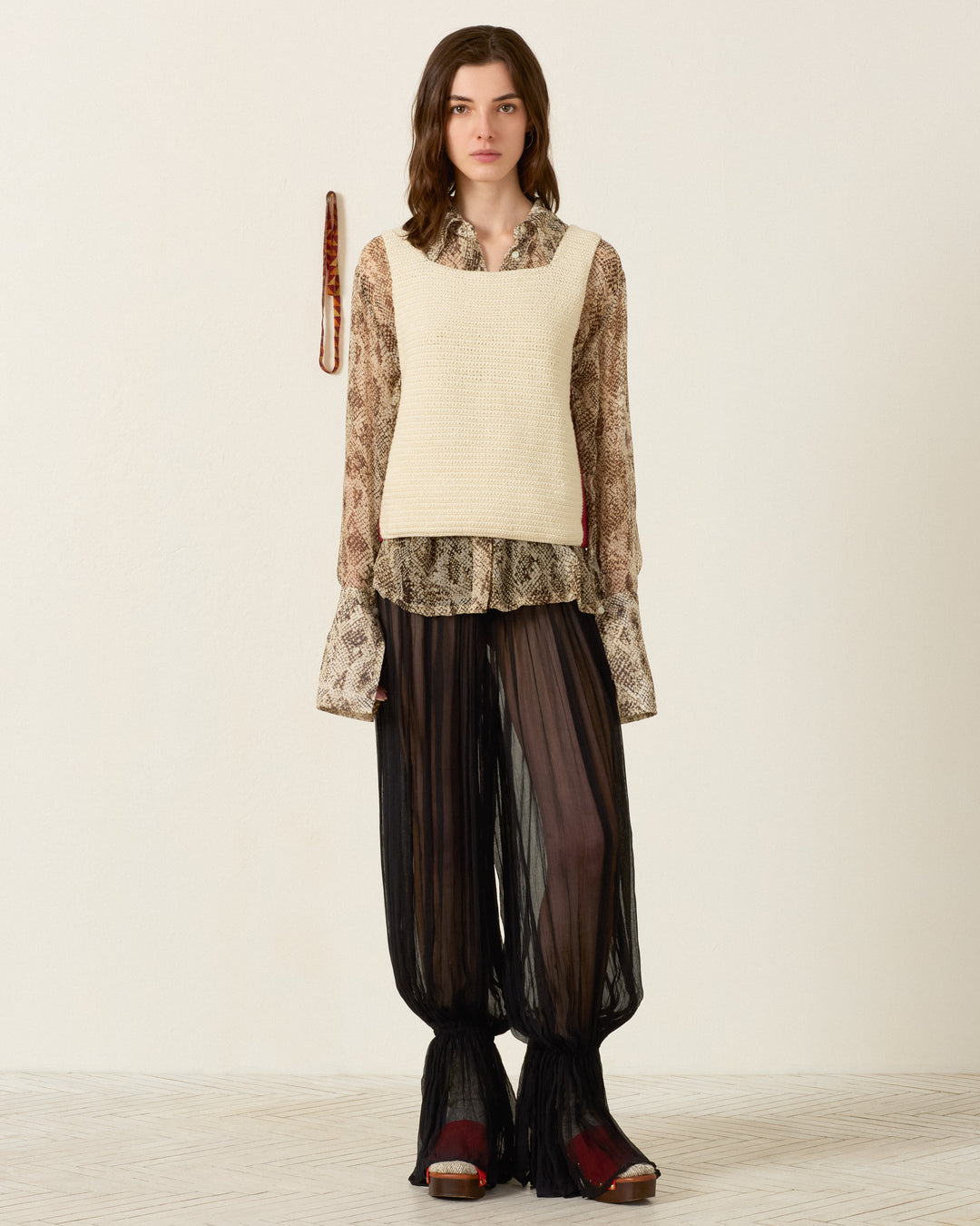 A woman stands against a plain wall, wearing a beige sleeveless top over a patterned shirt, the Keats Onyx Chiffon Pant—sheer black wide-legged harem pants with an elasticated waist in luxurious cupro-chiffon—and platform sandals.