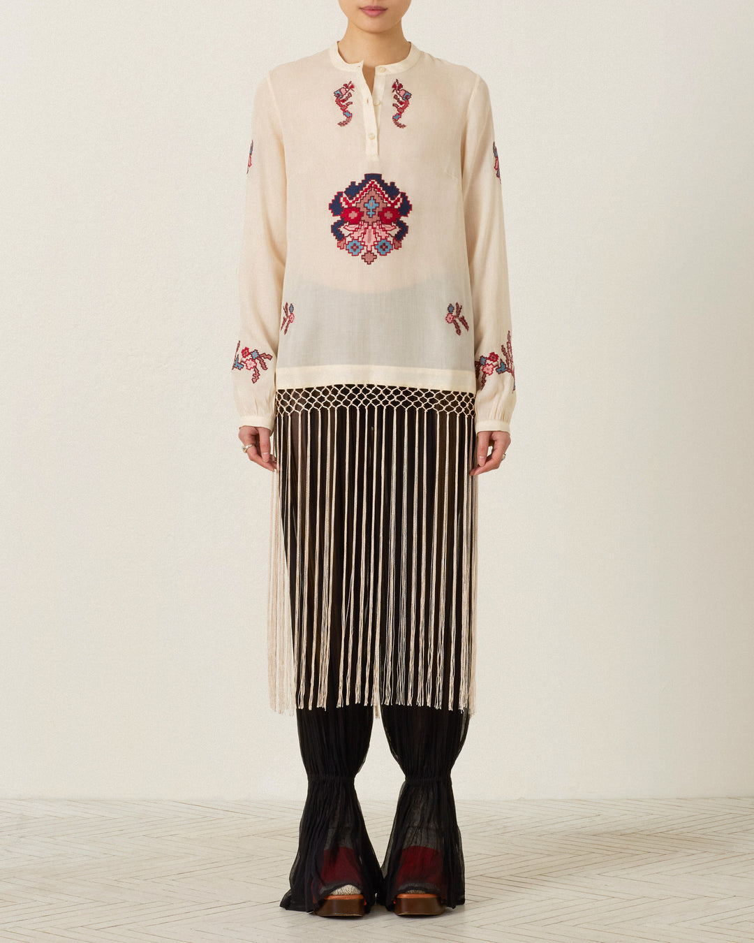 Wearing the Rune Kilim Stone Fringe Top—a sheer, long-sleeve design adorned with red hand-embroidered details and kilim motifs—paired with a black skirt featuring long cream fringe, black pants, and red-brown shoes, the person stands against a plain background.