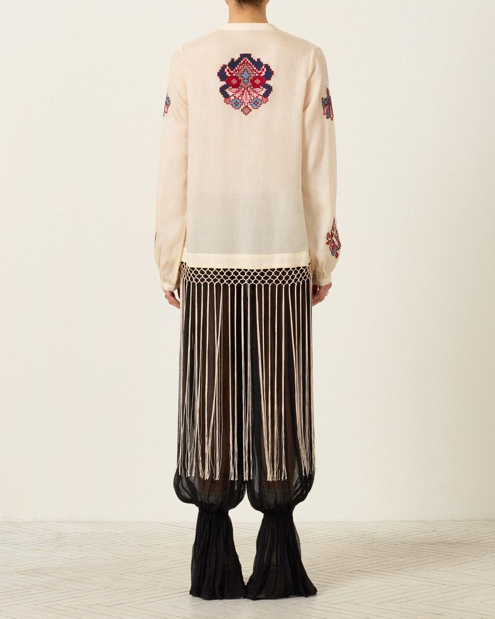 A person stands facing away, wearing the Rune Kilim Stone Fringe Top—a cream sheer blouse adorned with colorful hand-embroidered kilim motifs—paired with a long black skirt featuring vertical white fringe and a sheer hem, set against a plain light background.