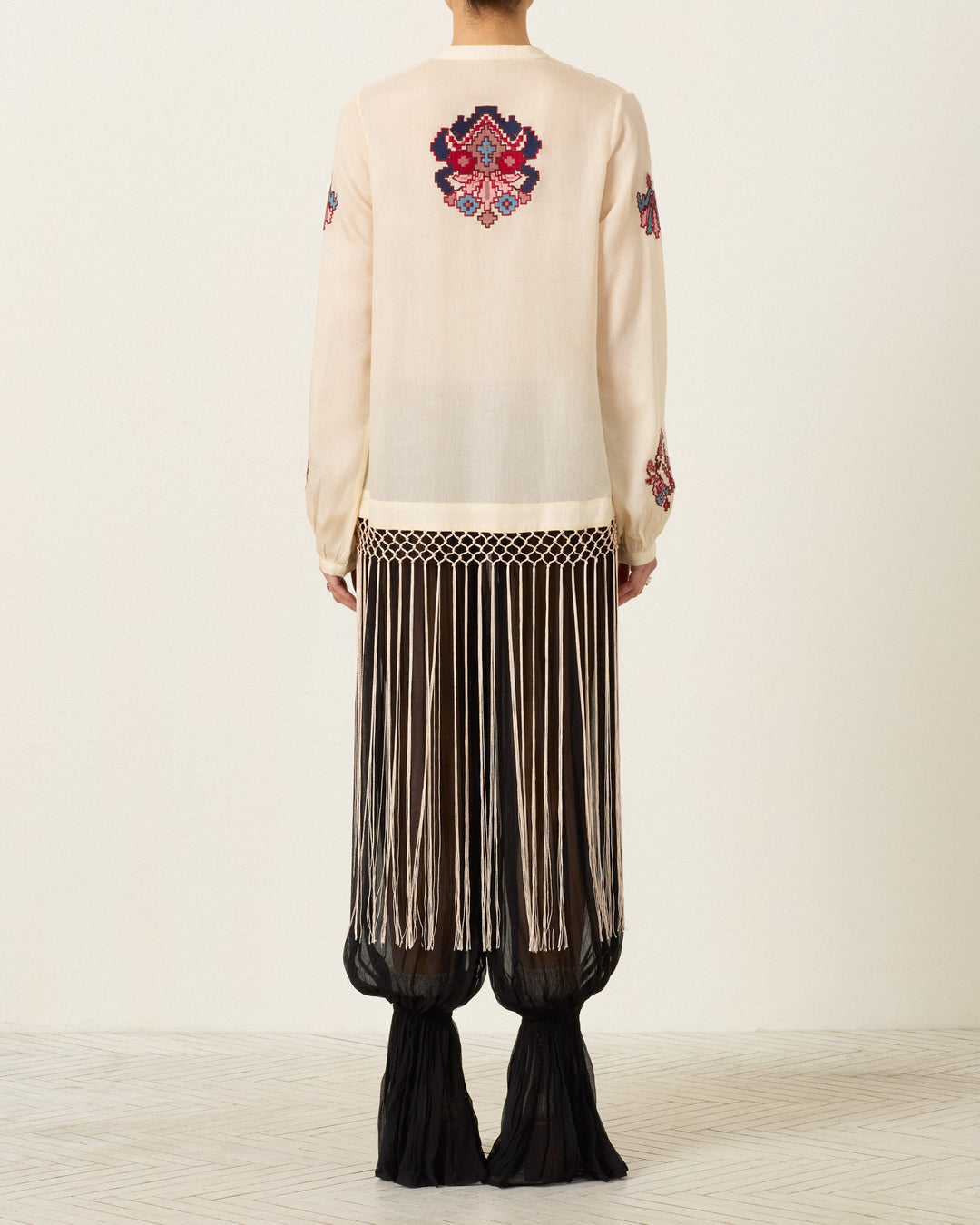 A person stands facing away, wearing the Rune Kilim Stone Fringe Top—a cream sheer blouse adorned with colorful hand-embroidered kilim motifs—paired with a long black skirt featuring vertical white fringe and a sheer hem, set against a plain light background.