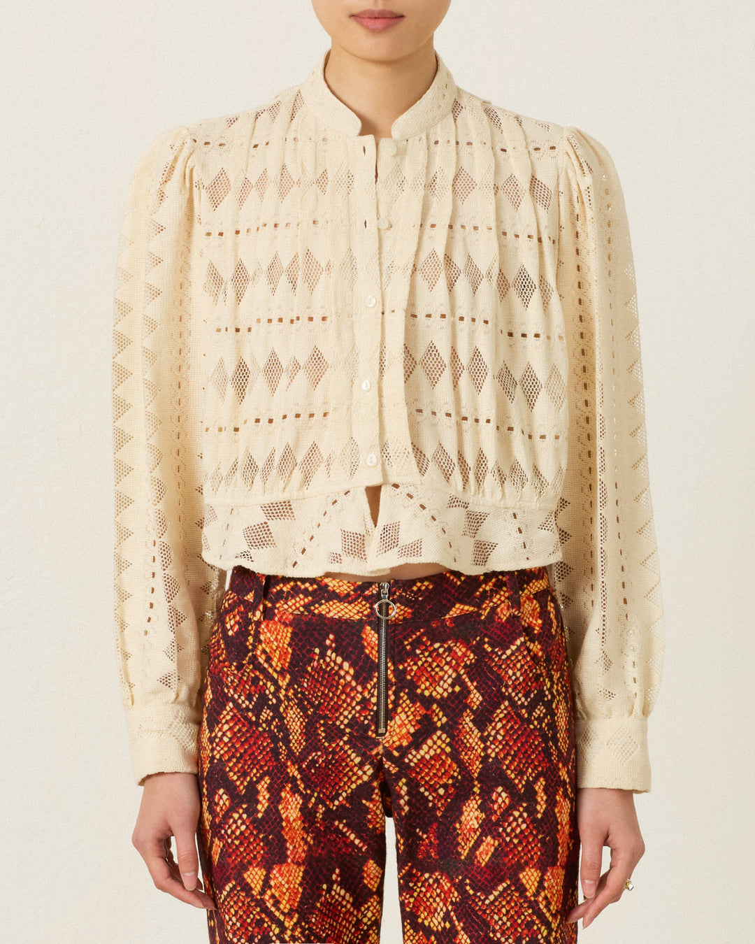 A person wears the Ryland Savanna Lattice Shirt, a cream, long-sleeve button-up blouse with geometric lace, paired with high-waisted, red and orange snakeskin-patterned pants.