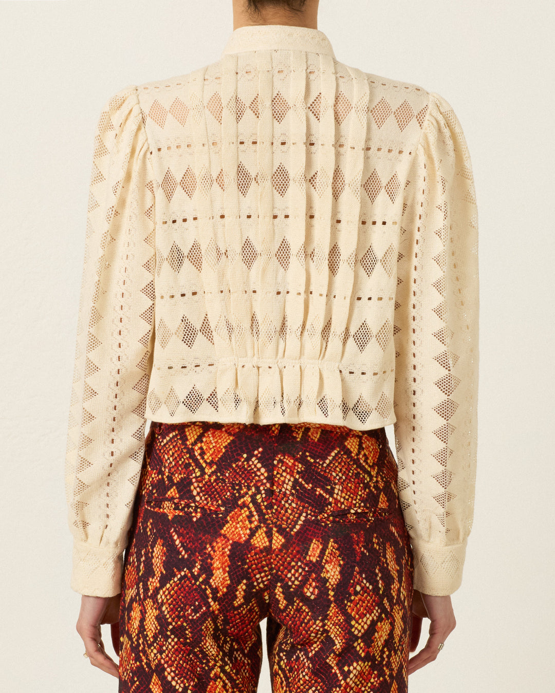 A person wearing the Ryland Savanna Lattice Shirt—a cream, long-sleeve blouse with a geometric lace design and Victorian silhouette—paired with red snake-print pants, shown from the back against a plain background.