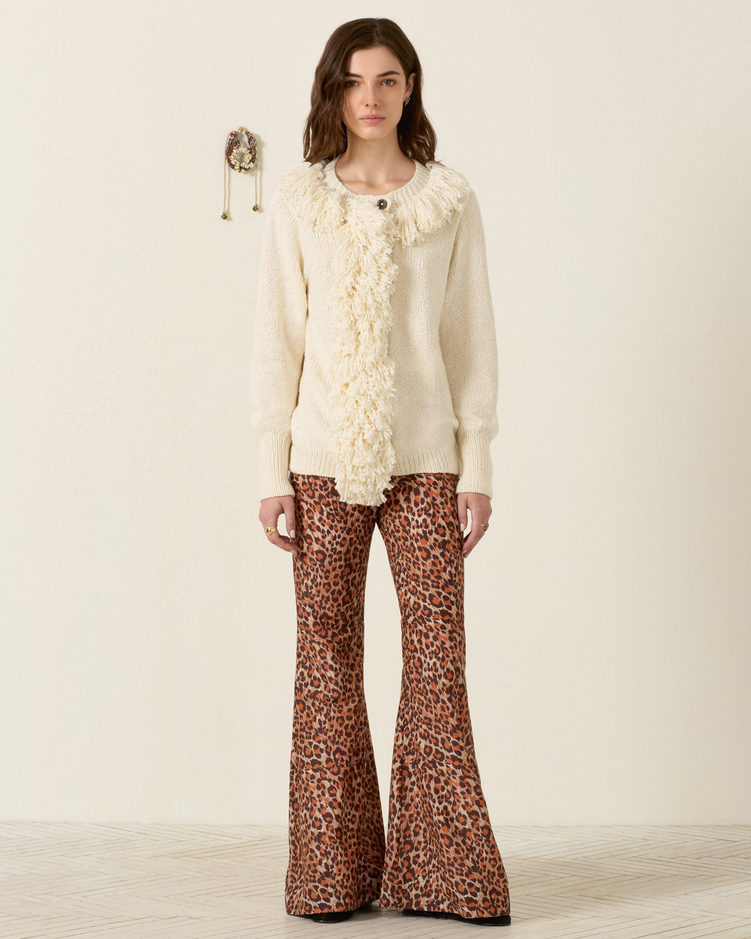 A person stands indoors against a plain wall, wearing the Sammi Bone Fringe Cardigan—a cream artisan-made sweater with textured detailing—paired with brown leopard print flared pants.