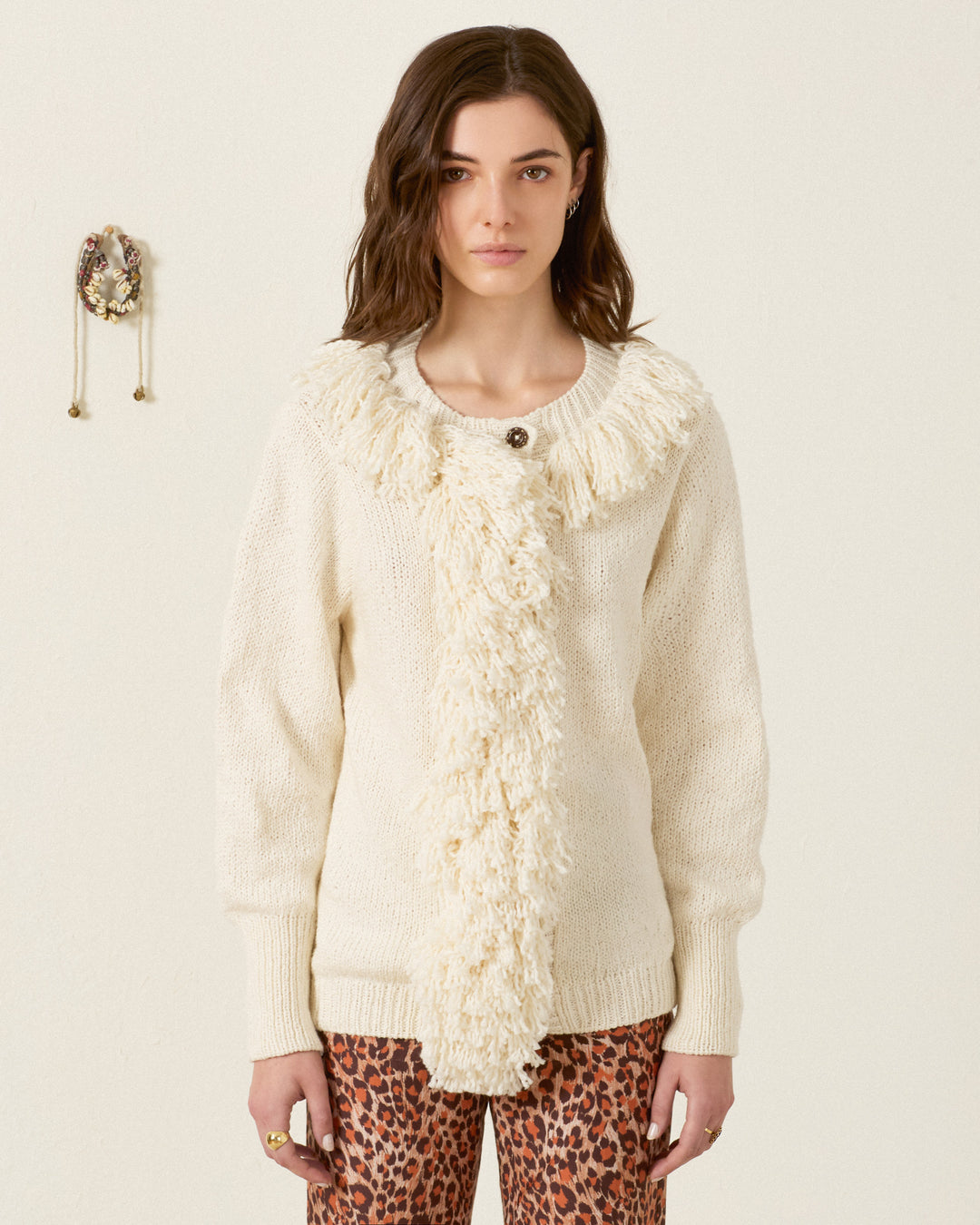 Woman wearing the Sammi Bone Fringe Cardigan, an artisan-made cream sweater featuring textured fringe detail down the front and collar, paired with leopard print pants and standing against a plain indoor background.