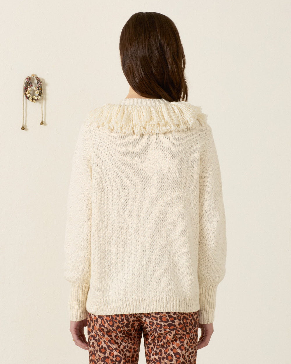 A person with long brown hair is seen from behind against a plain light wall, wearing the artisan-made Sammi Bone Fringe Cardigan paired with leopard print pants.