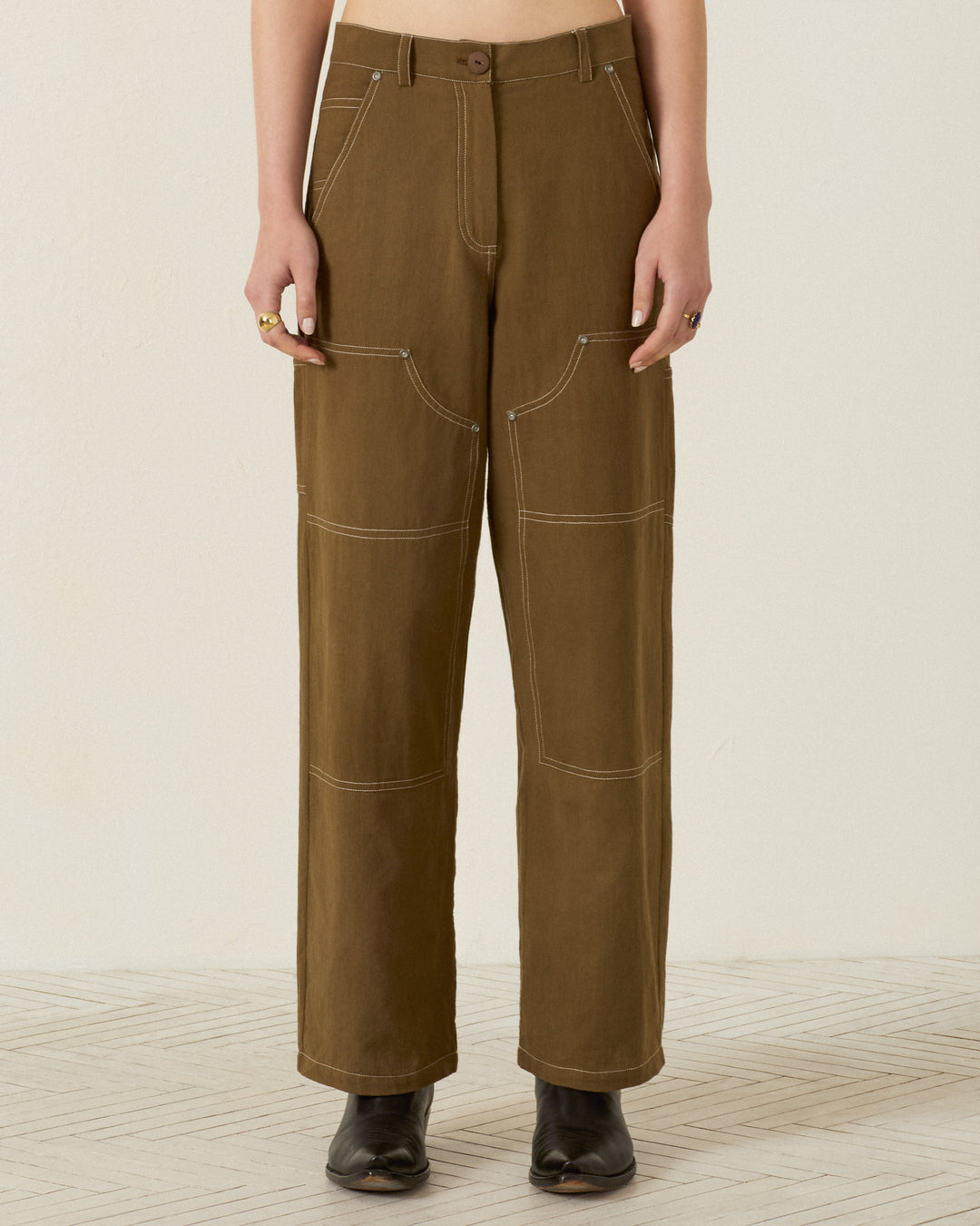 Wearing the Scottie Olive Branch Pant—high-waisted, olive-green utility workwear pants made from khadi cotton with white contrast stitching and large front pockets—paired with black boots.