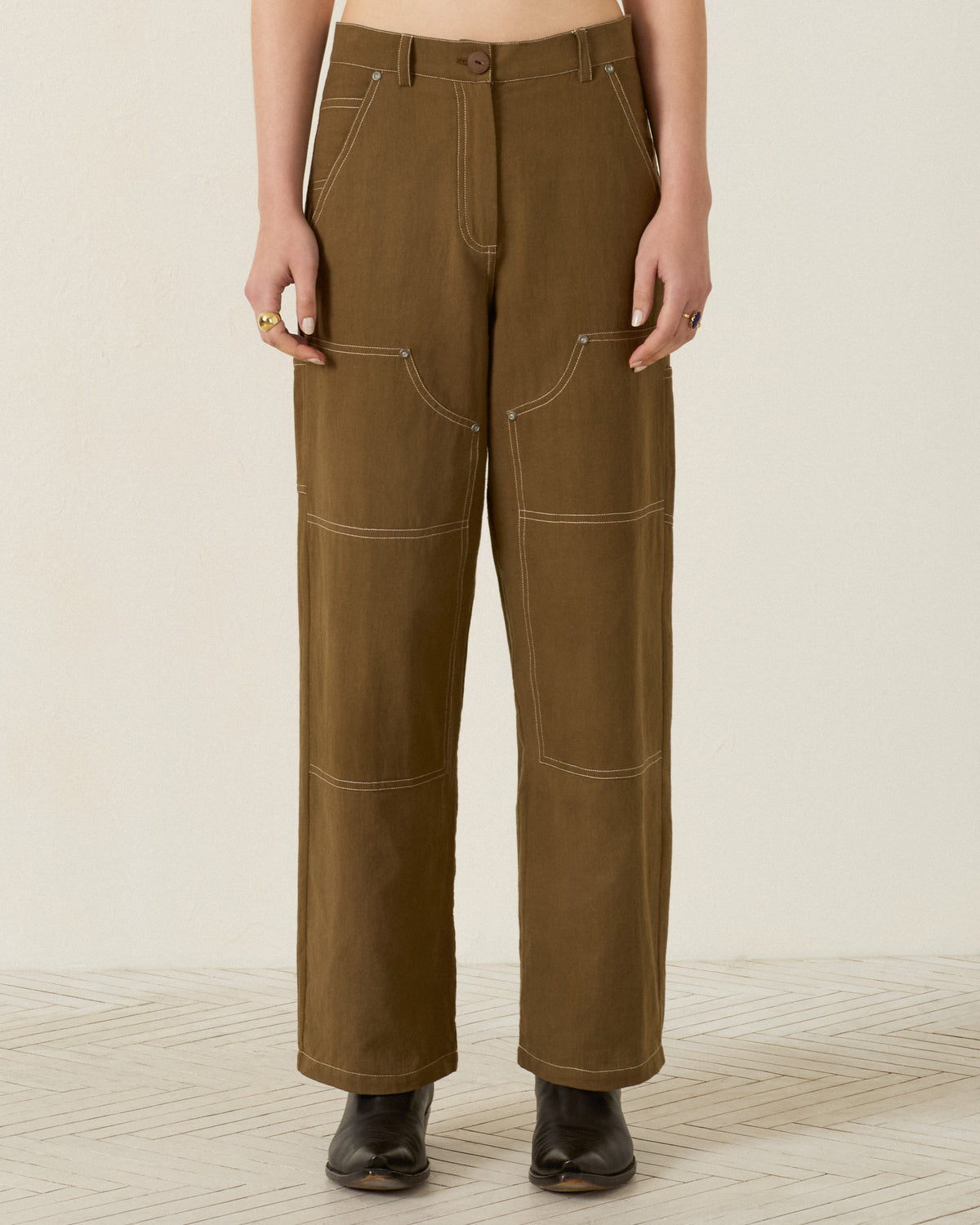 Wearing the Scottie Olive Branch Pant—high-waisted, olive-green utility workwear pants made from khadi cotton with white contrast stitching and large front pockets—paired with black boots.
