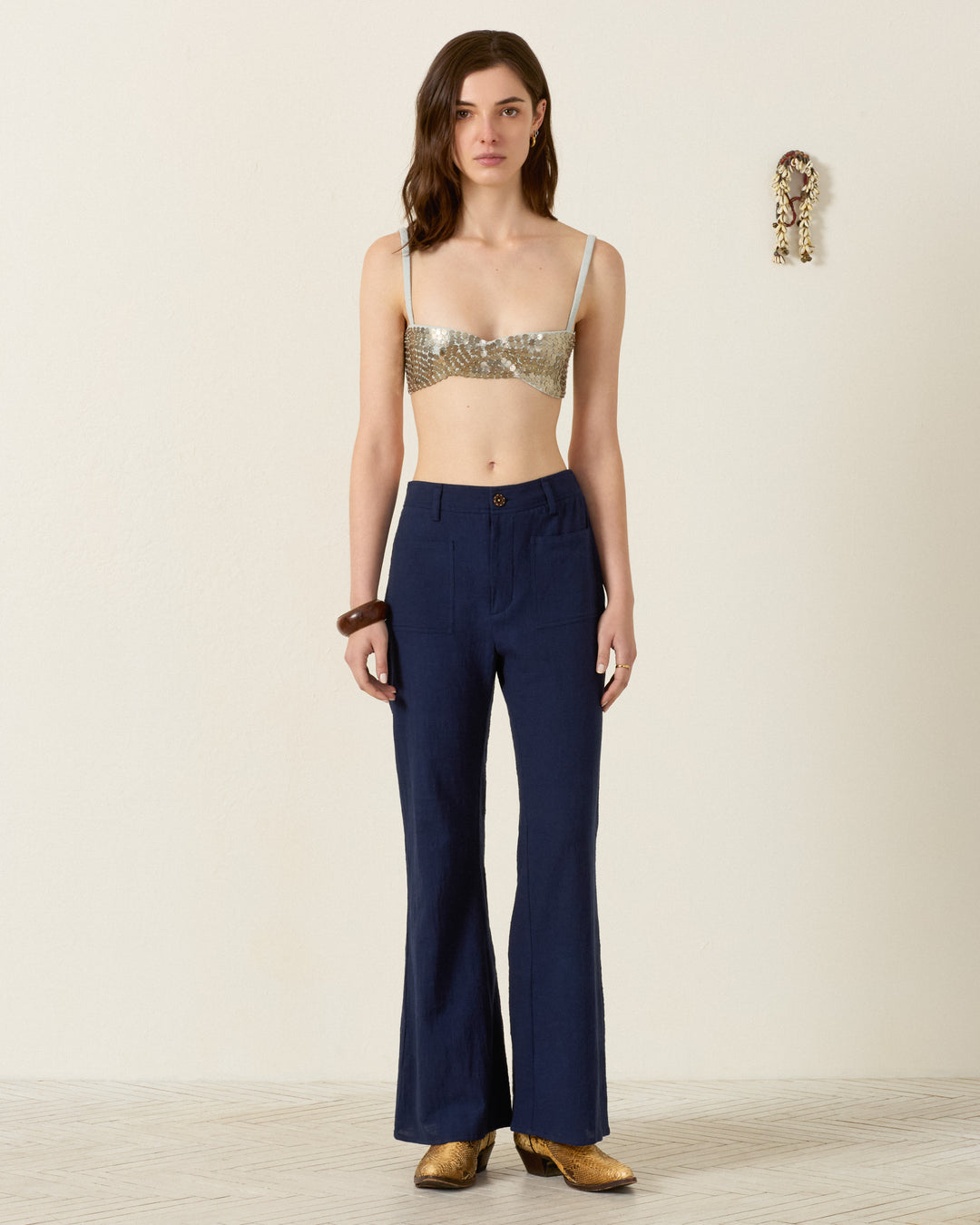 A woman stands indoors wearing a gold sequin bralette, gold shoes, a large bangle, and the Sonnet Indigo Night Jean—navy flared-leg pants with an elasticated waistband—with a small wall decoration visible in the background.