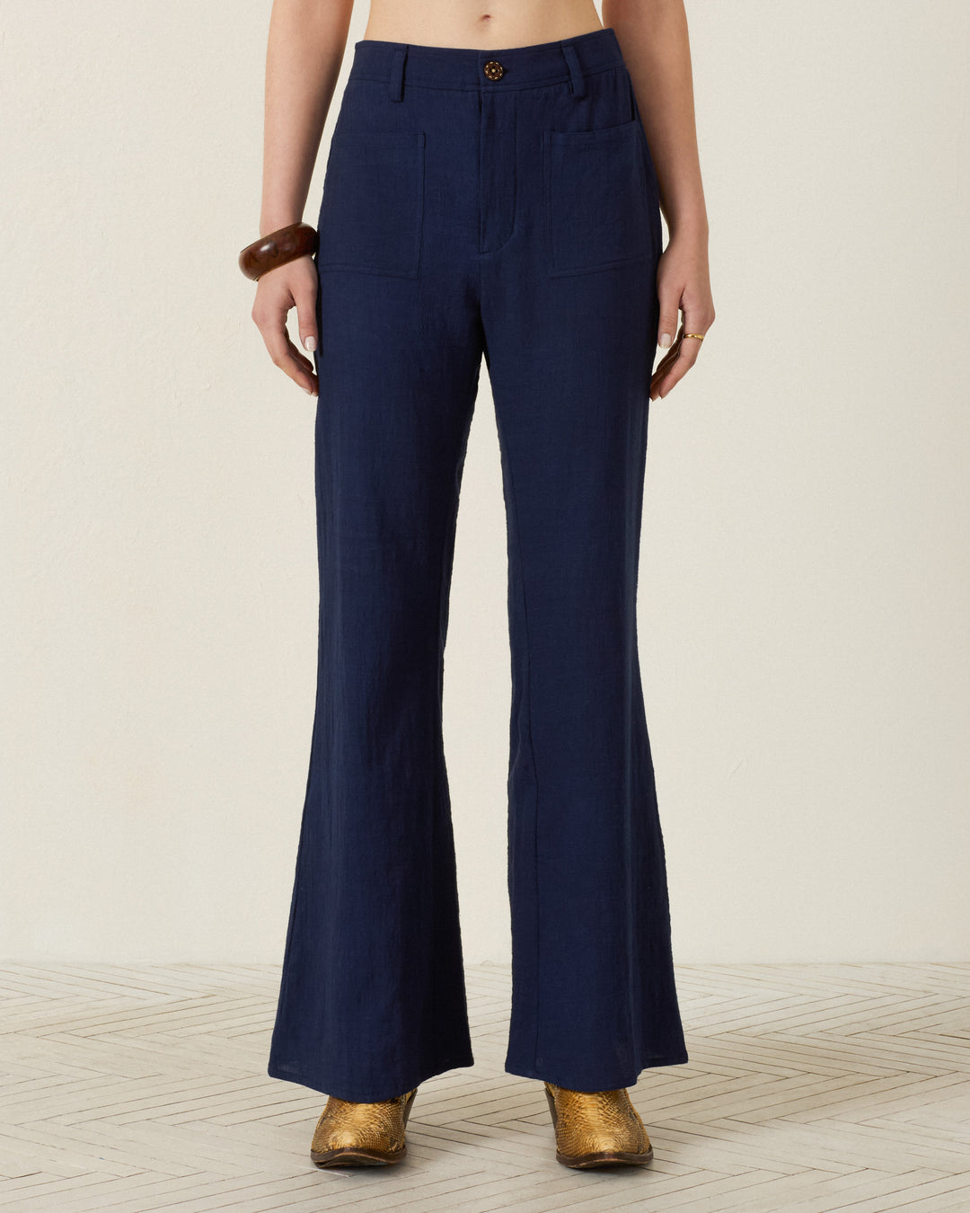 Wearing the Sonnet Indigo Night Jean—high-waisted, navy blue flare pants with an elasticated waistband and front pockets—styled with gold shoes, a dark bracelet, and rings, standing against a plain light-colored background.