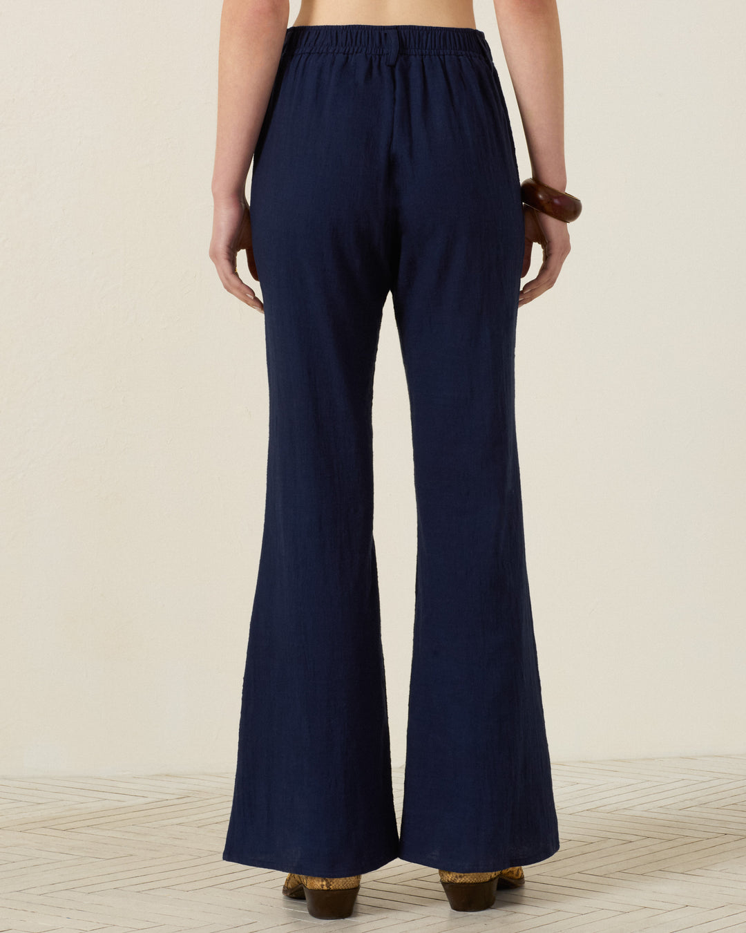 A person standing on a light patterned floor, wearing the Sonnet Indigo Night Jean—navy blue, high-waisted, wide-leg pants with an elasticated waistband—and brown shoes, photographed from the back.