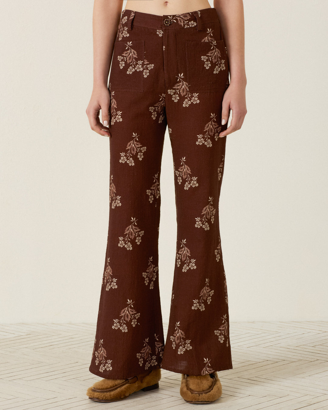 A person wearing the Sonnet Doe Elderflower Jean—high-waisted, brown cotton pants with a floral print and flared leg—paired with tan moccasin shoes, standing on a light patterned floor.