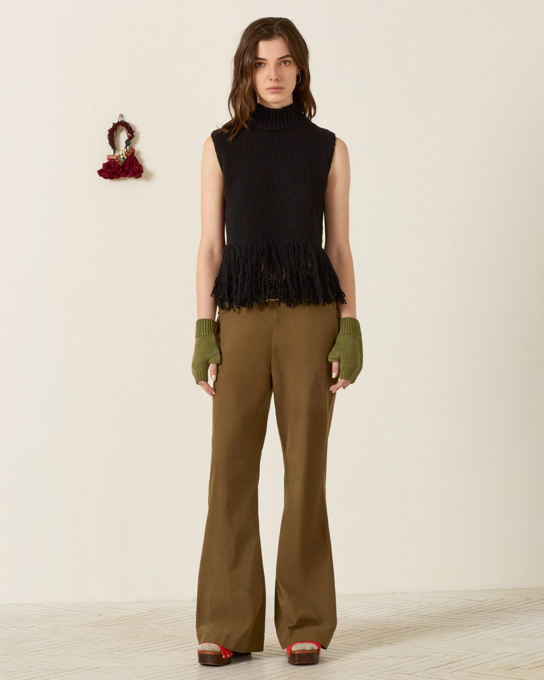 A woman stands against a plain wall wearing a sleeveless black turtleneck top, green fingerless gloves, red shoes, and the Charlie Olive Branch Pant—high-waisted pants crafted from khadi cotton in an army green hue. A small mirror hangs on the wall beside her.