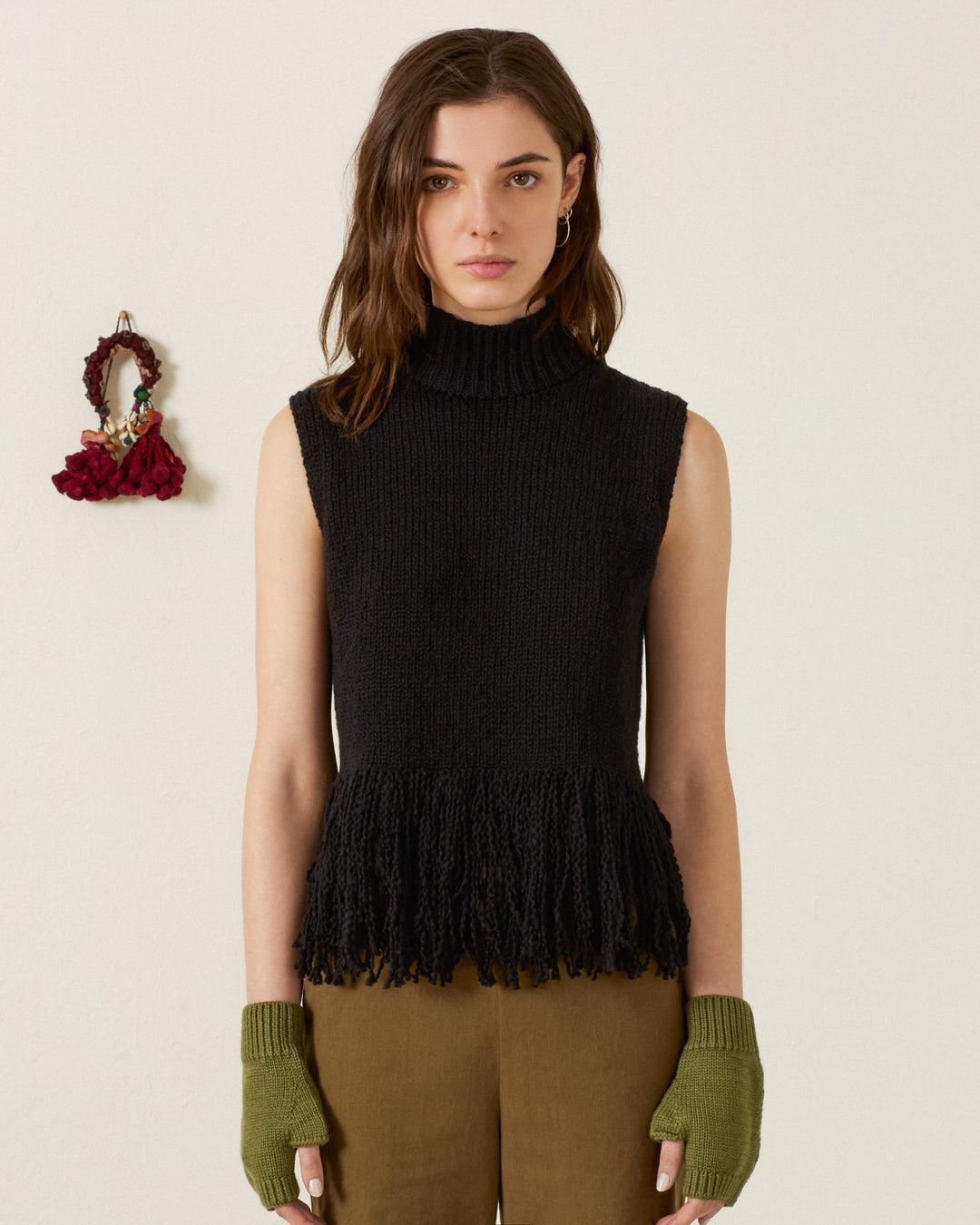 A woman stands against a plain wall wearing the Sonia Onyx Fringe Top—an artisan-made, sleeveless black hand-knitted top crafted from Peruvian cotton—paired with olive green fingerless gloves and brown pants, with a small decorative item hanging on the wall beside her.