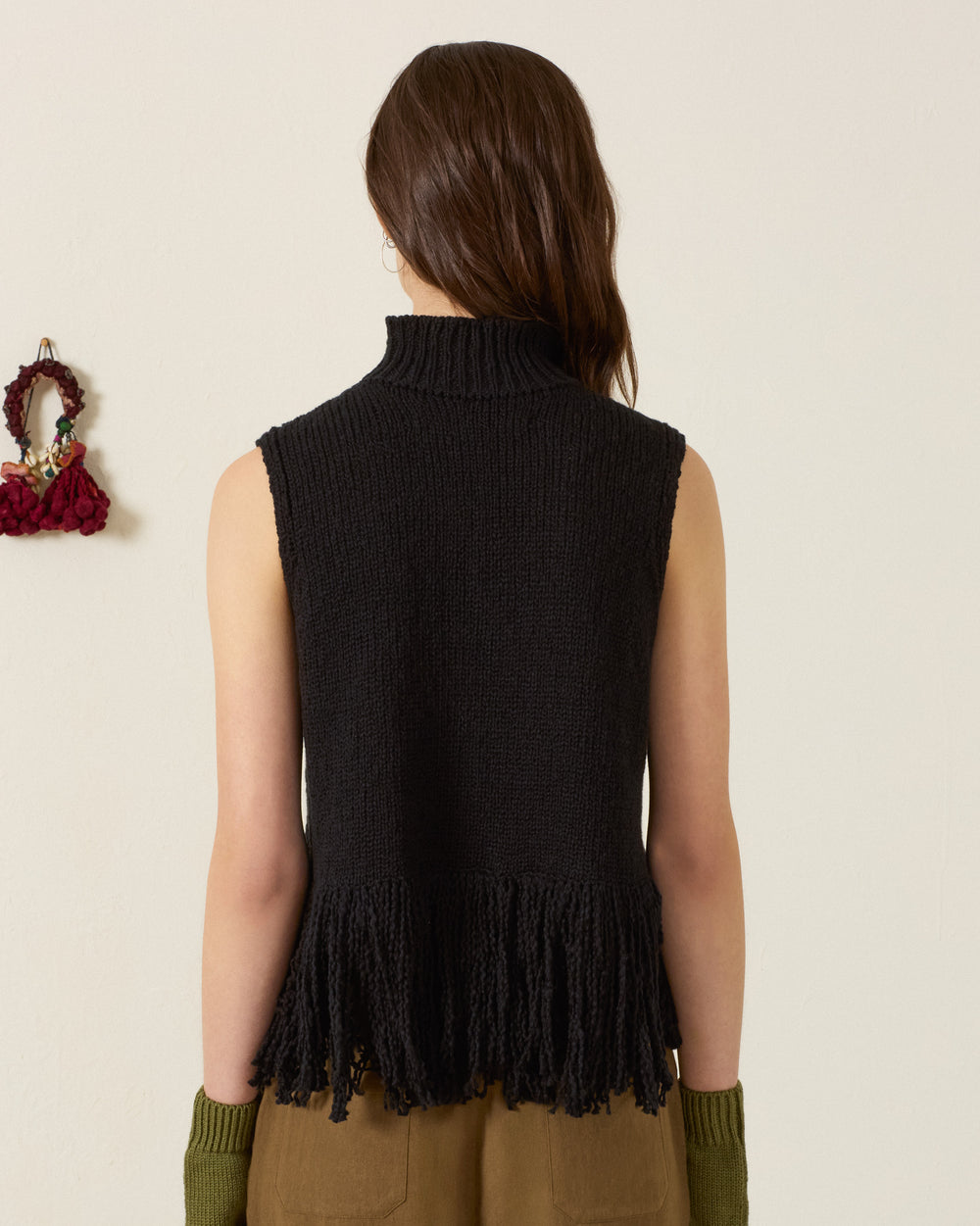A person with long brown hair, shown from the back, is wearing the sleeveless black hand-knitted Sonia Onyx Fringe Top with fringe at the hem and olive green arm warmers.