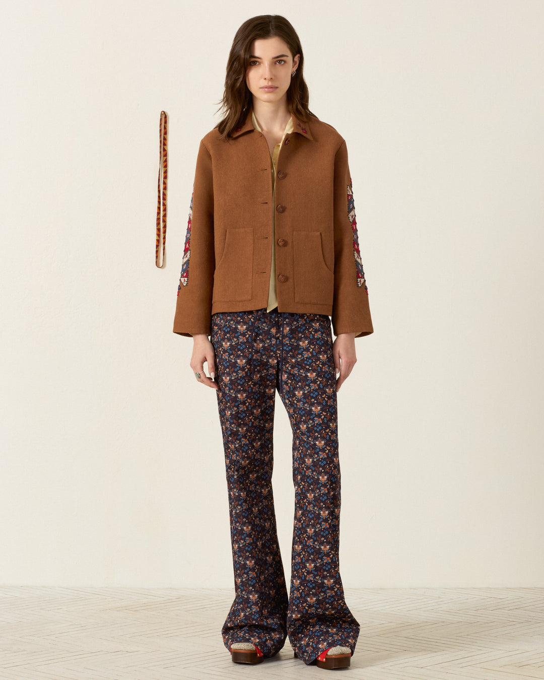 A person stands facing forward wearing the Western Doe Arrow Silk Jacket—a brown artisan piece with floral sleeves featuring delicate hand embroidery—paired with patterned flared pants and platform shoes against a plain background with a hanging accessory on the wall.