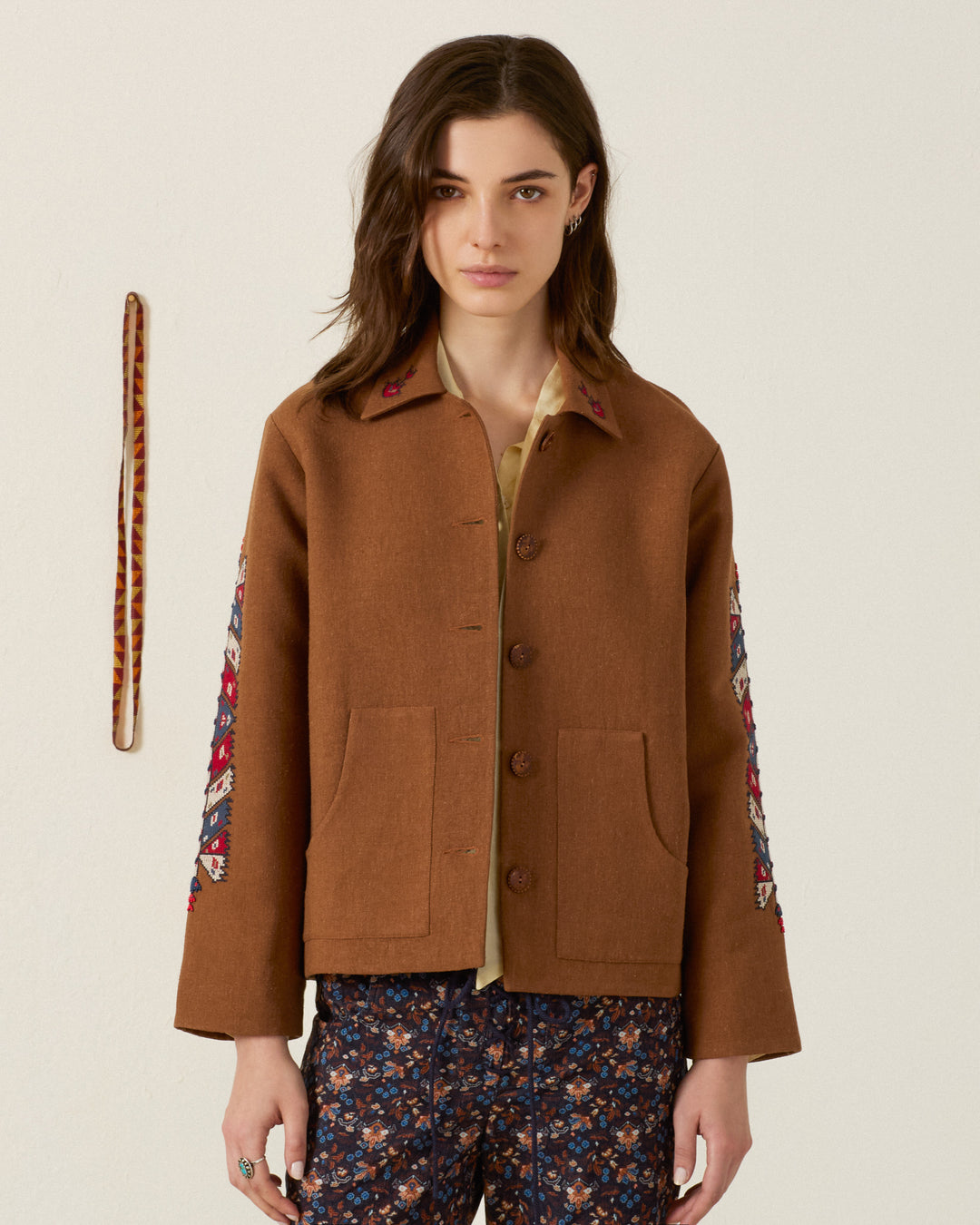 A woman wearing the Western Doe Arrow Silk Jacket, featuring brown artisan fabric and floral hand embroidery on the sleeves, stands against a plain background with a striped belt hanging on the wall.