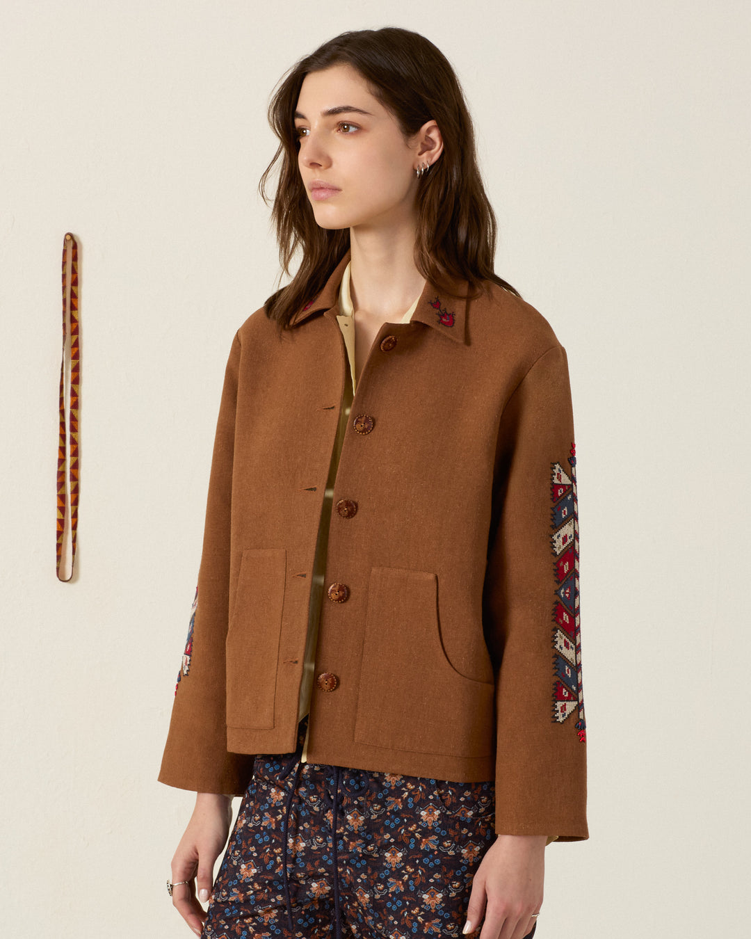 Dressed in the Western Doe Arrow Silk Jacket—featuring a rich brown hue, patterned sleeves, and intricate hand embroidery—a person pairs it with floral pants while standing against a plain wall adorned with a hanging woven strip in the background.