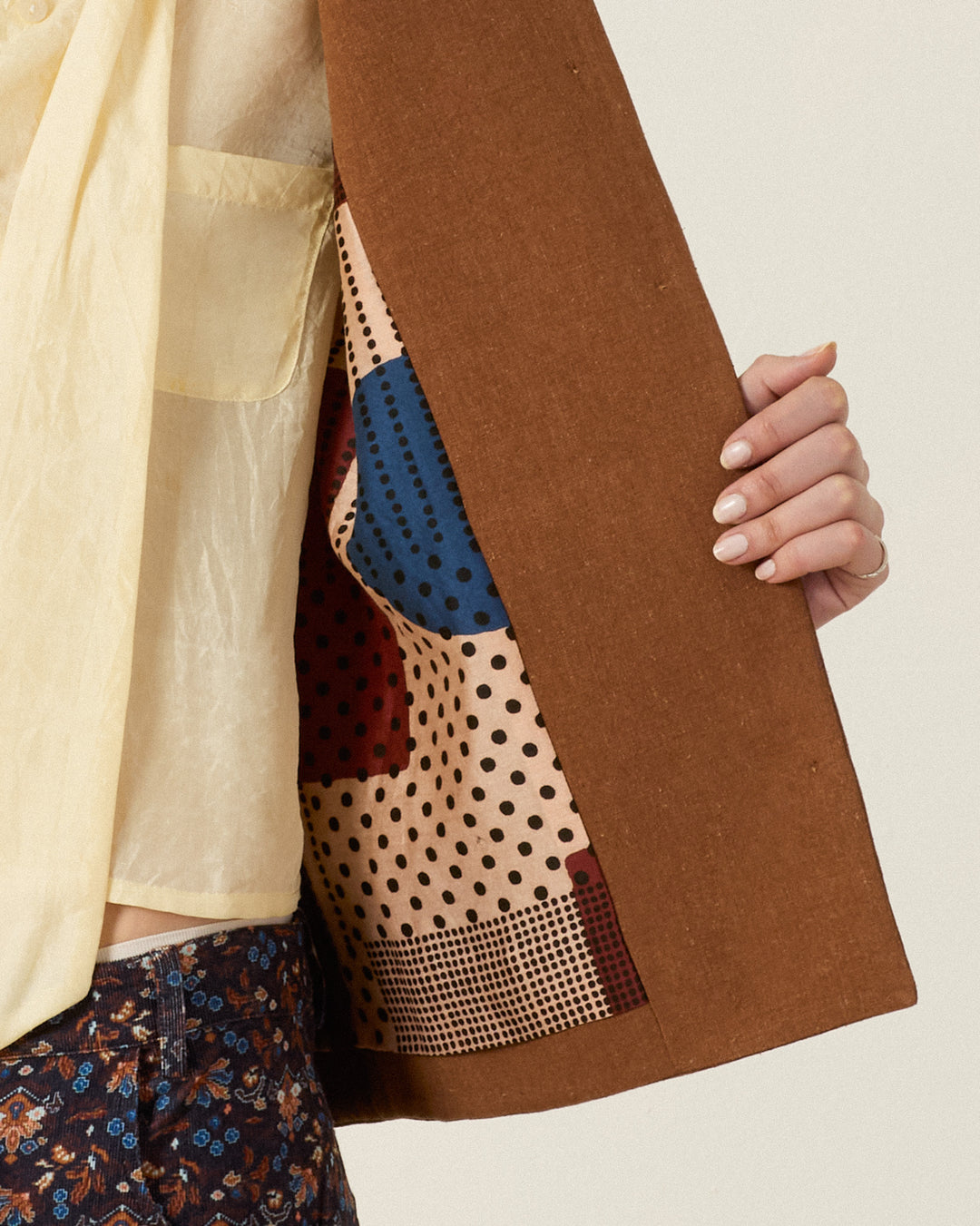 A person holds open the Western Doe Arrow Silk Jacket, showcasing its patterned lining with dots and geometric shapes, worn over a yellow shirt and floral pants.