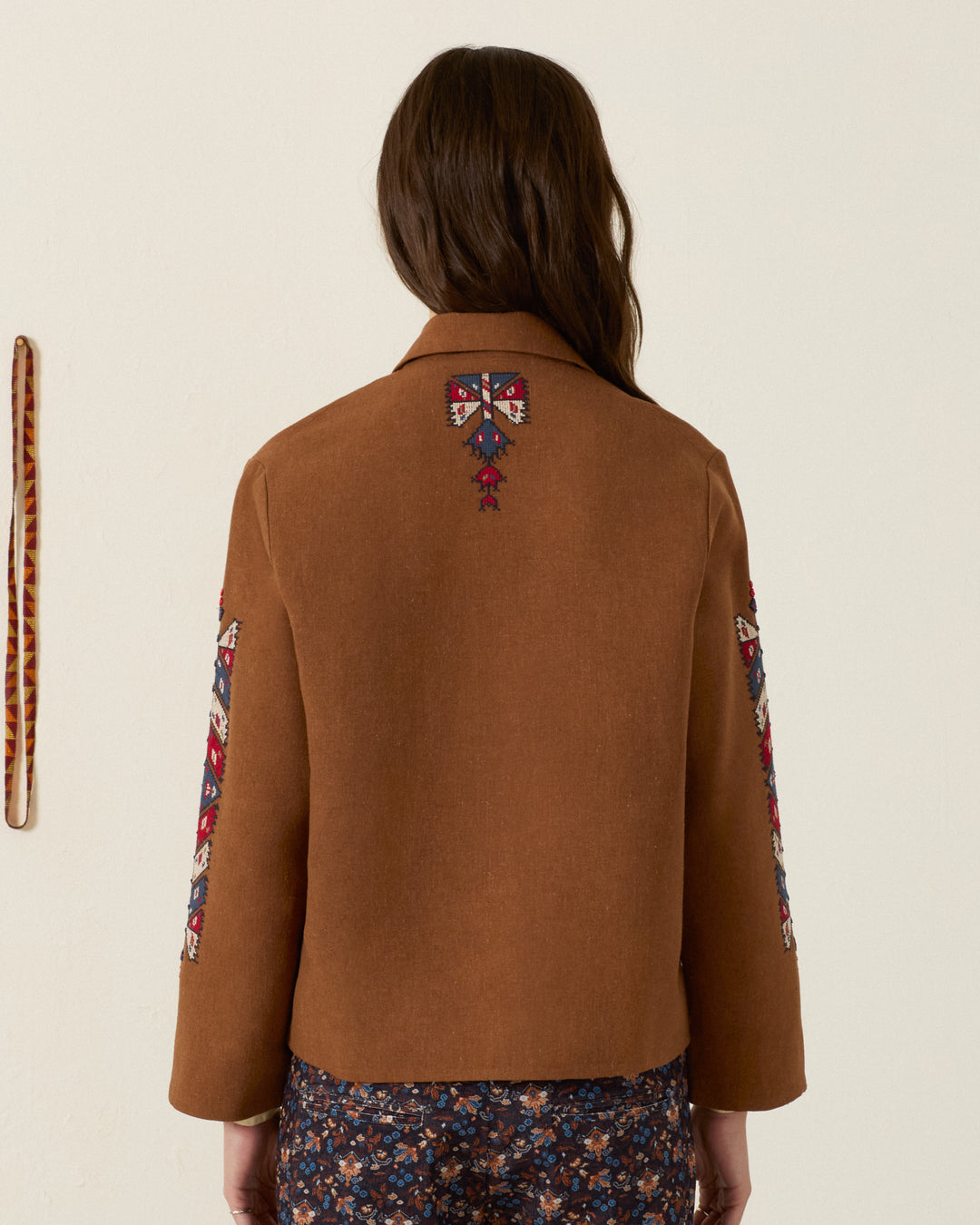 A person with long brown hair wears the Western Doe Arrow Silk Jacket—an elegant artisan piece featuring hand embroidery on the back and sleeves—while standing against a plain wall.