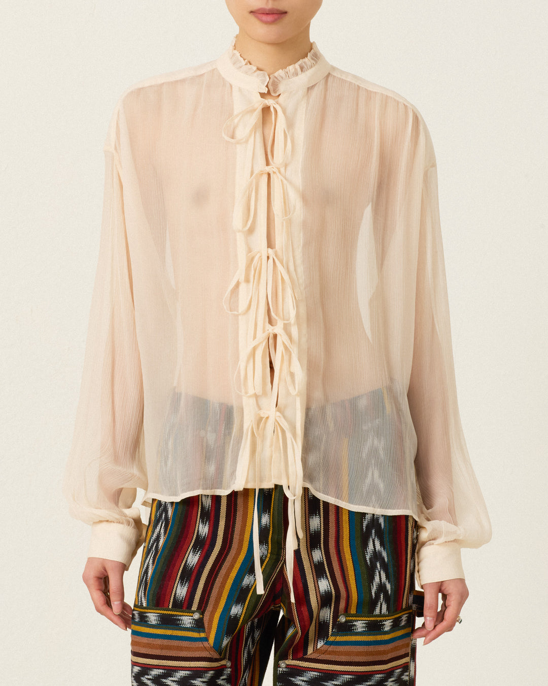 A person wearing the Willa Prairie Tint Shirt—a sheer, long-sleeved cream blouse with a ruffled collar and multiple front ties, crafted from delicate cupro-chiffon—paired with colorful patterned pants, standing against a plain background.