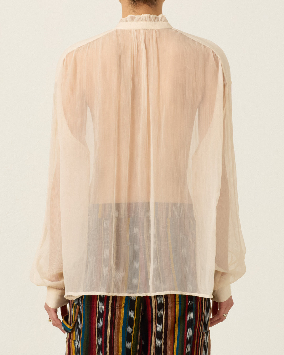 A person photographed from the back against a plain background is wearing the Willa Prairie Tint Shirt, a sheer cupro-chiffon, long-sleeved, light-colored blouse with a ruffled collar, paired with a multicolored striped skirt.