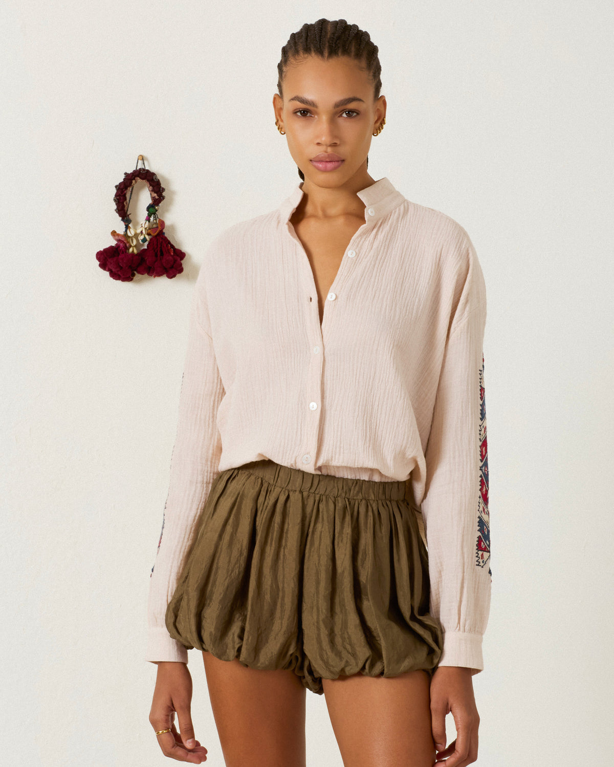 A woman stands against a white wall, wearing the Kiki Prairie Arrow Shirt—a light pink cotton crepe shirt with an Edwardian silhouette—paired with olive green high-waisted shorts; a decorative item hangs on the wall beside her.