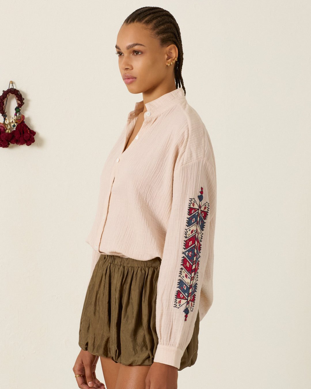 A woman stands in profile against a plain wall, wearing the Kiki Prairie Arrow Shirt—a light, long-sleeve cotton crepe top featuring an elegant embroidered pattern on the sleeve—paired with olive green shorts.