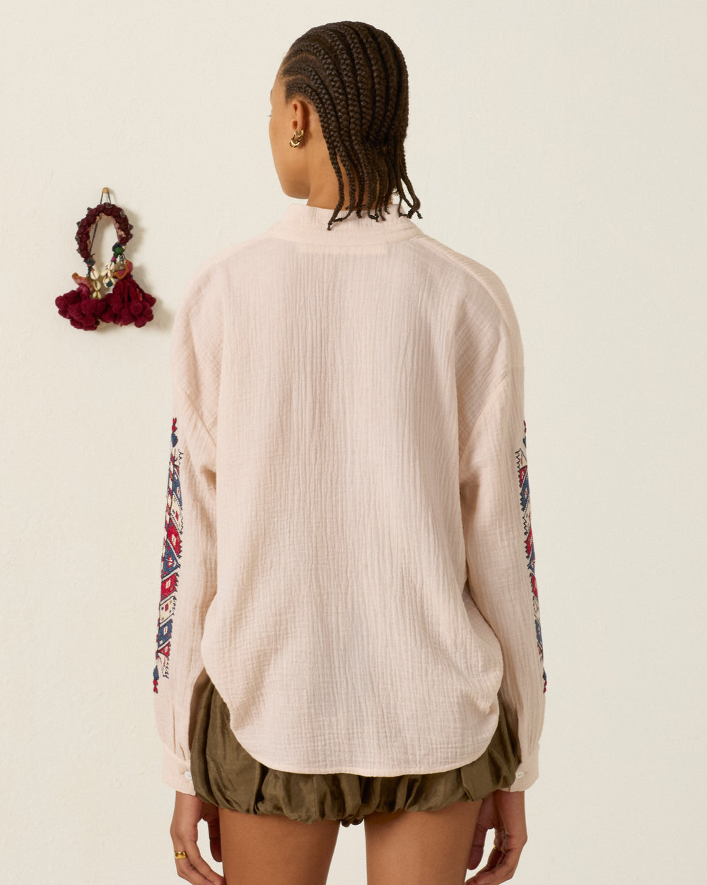 A person with braided hair, seen from the back, is wearing the Kiki Prairie Arrow Shirt—a light cotton crepe top featuring hand embroidery on the sleeves—paired with olive shorts and standing in front of a white wall adorned with a maroon hair accessory.