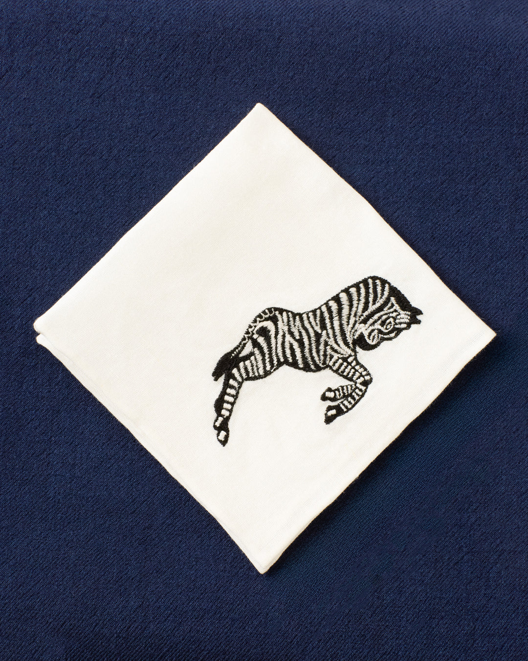 The Cai Zebra Canter Handkerchief, a white linen-cotton accessory embroidered with a zebra motif, is displayed on dark blue textured fabric, making it a charming good luck gift.