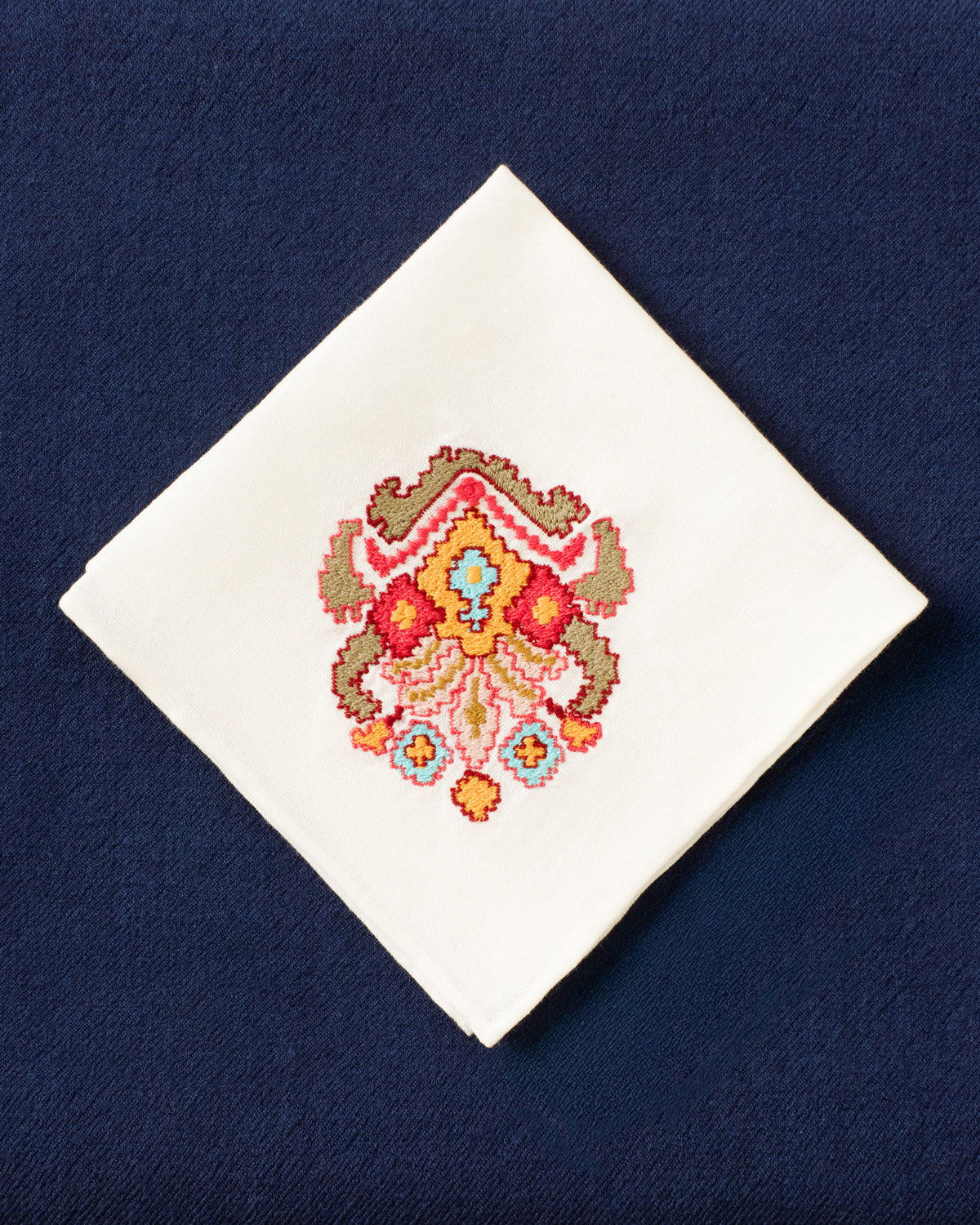 The Cai Kilim Emblem Handkerchief, featuring a colorful embroidered floral pattern on white linen-cotton fabric, is displayed on a dark blue textured surface.