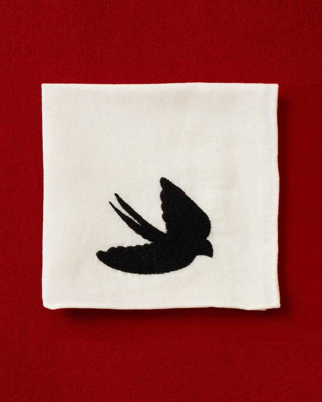 The Cai Swallow Swoop Handkerchief, made from white linen-cotton and featuring a black silhouette of a flying swallow, is displayed against a red textured background.