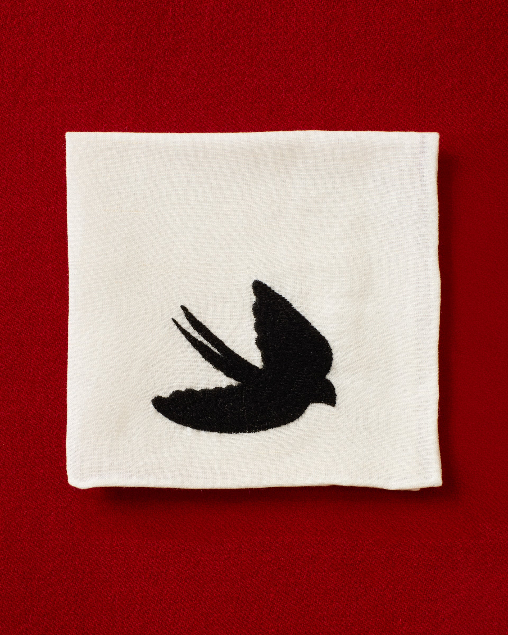The Cai Swallow Swoop Handkerchief, made from white linen-cotton and featuring a black silhouette of a flying swallow, is displayed against a red textured background.