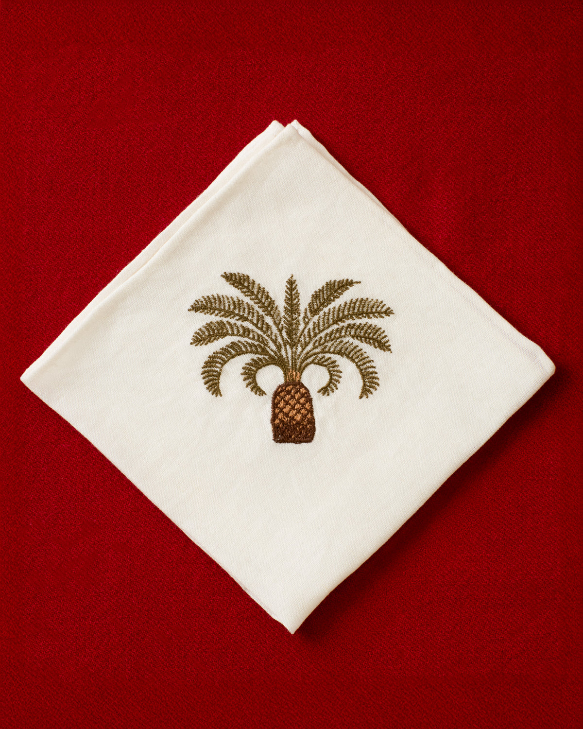 The Cai Palm Branch Handkerchief, crafted from white linen-cotton and featuring an embroidered brown palm tree motif, is displayed on a red textured surface.