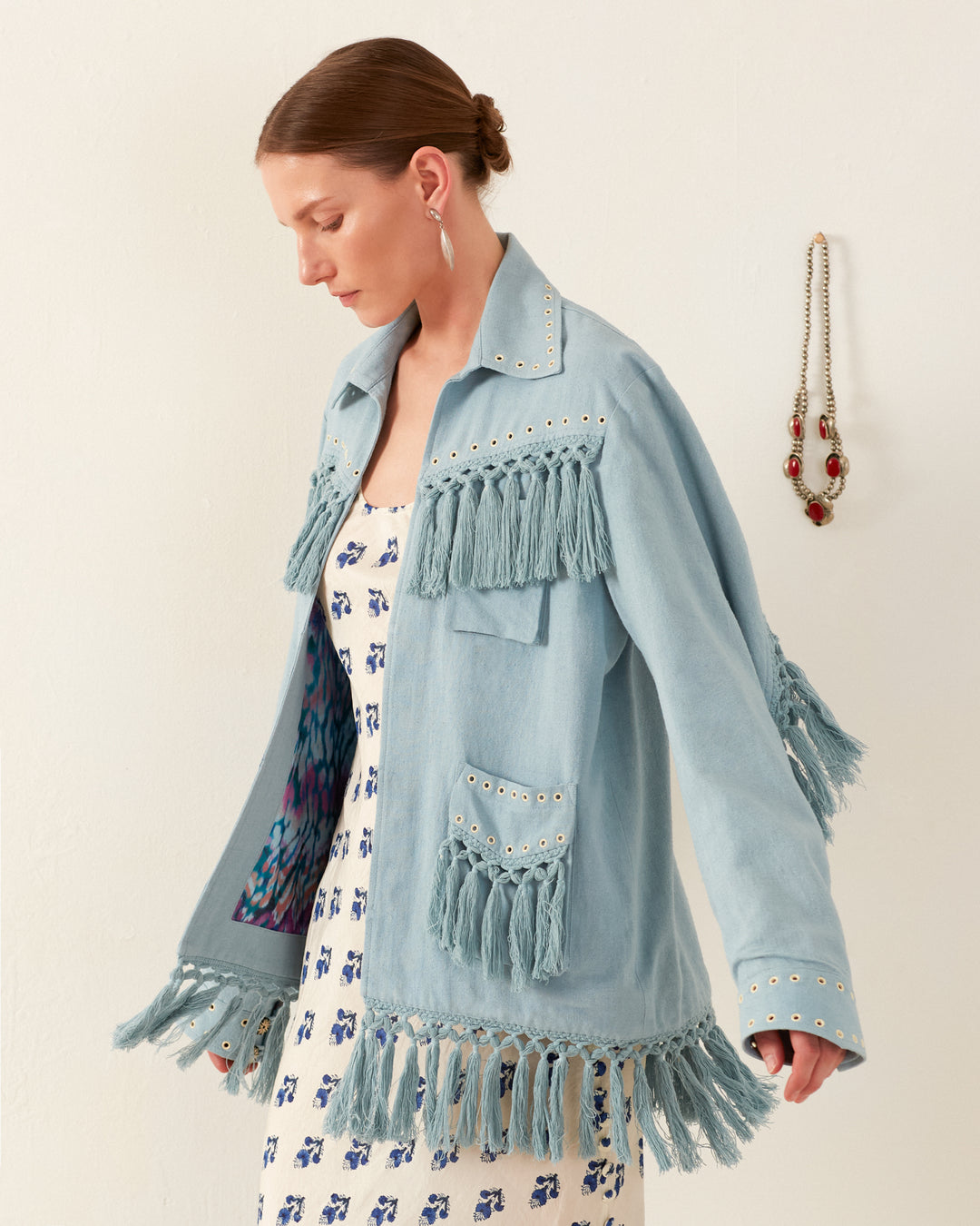 A person wearing the Jude Aqua Fringe Jacket, featuring silver eyelets, over a white dress with blue patterns. They have short brown hair and are accessorized with hoop earrings. In the background, a hand-embellished necklace hangs on the wall, adding an artistic touch to the scene.