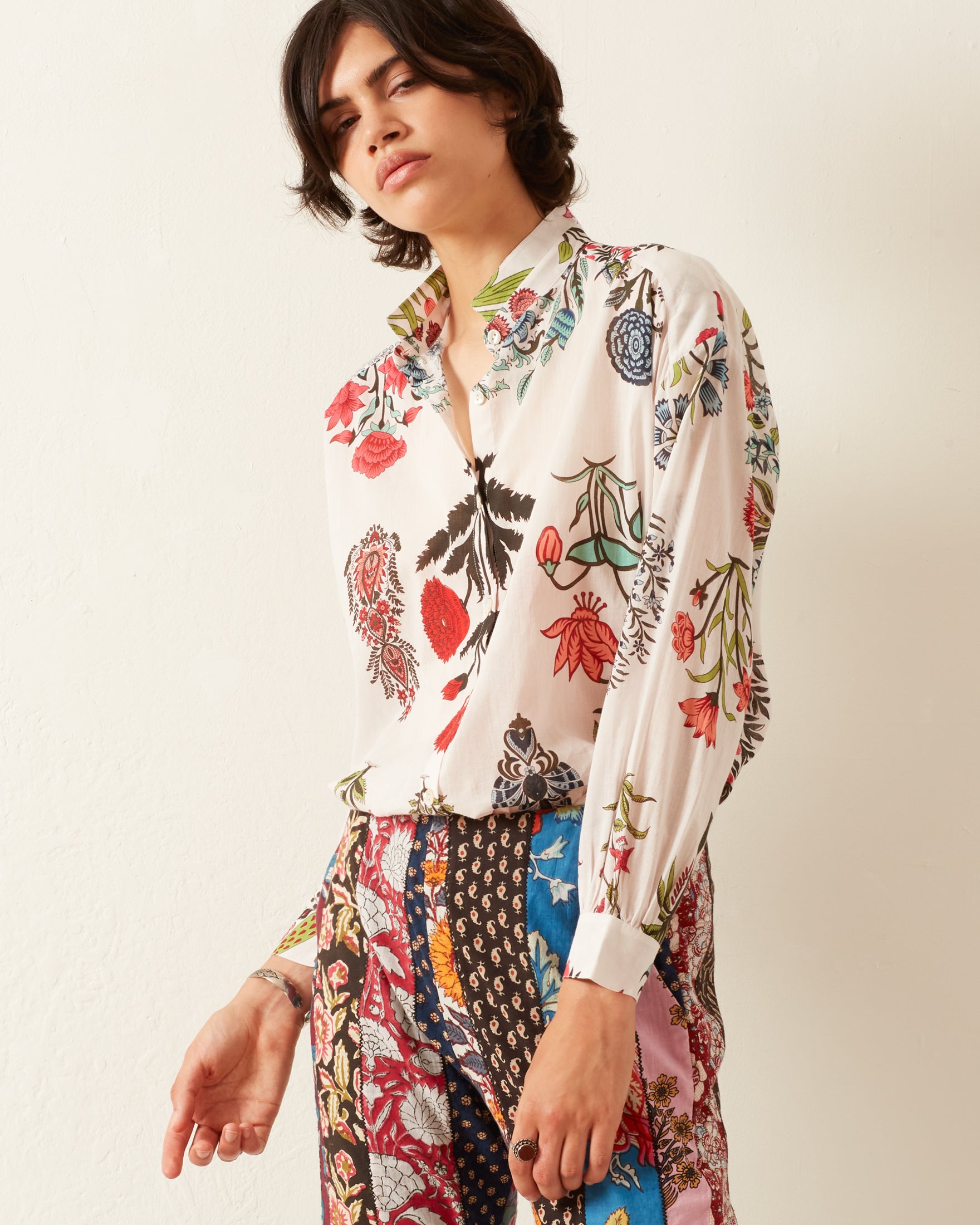 A person wearing the Kiki Flower Show Shirt, featuring Mother of Pearl buttons and paired with patchwork pants, stands against a plain background, gazing directly at the camera. The ensemble beautifully combines vibrant botanical and geometric designs, crafted with traditional hand-printed cotton techniques.