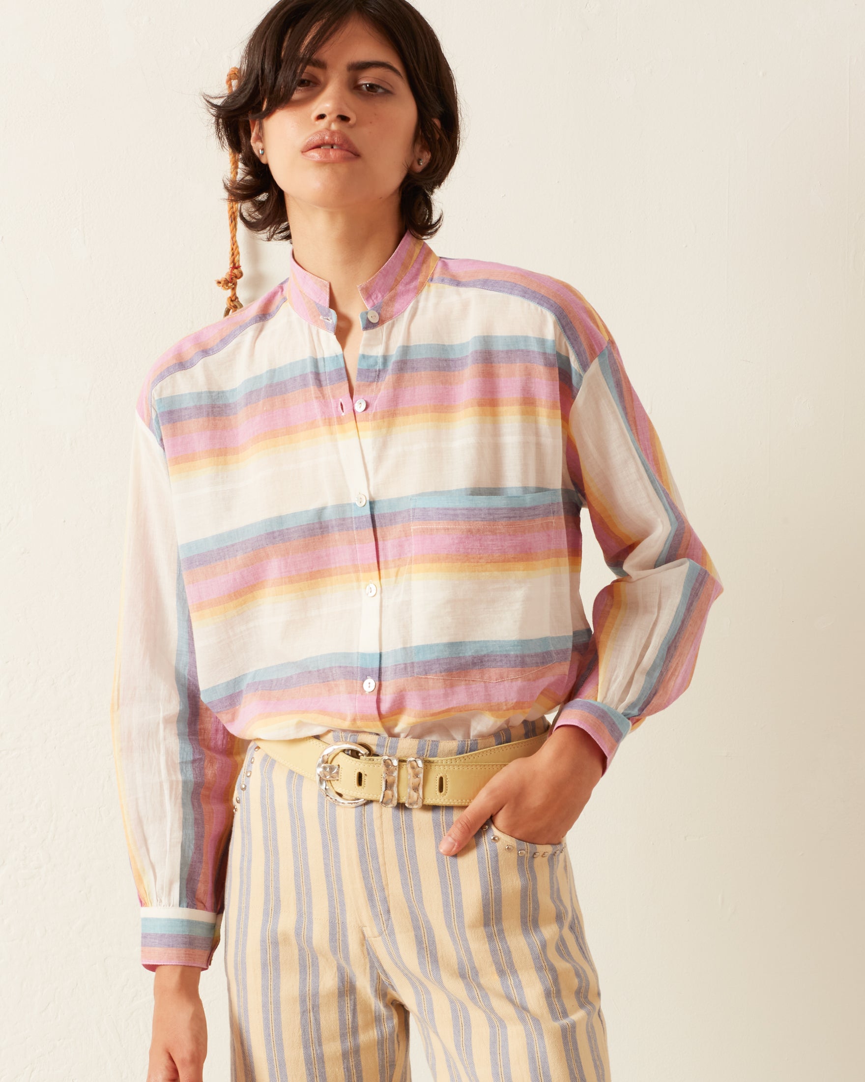 A person wearing the Kiki Sunset Shirt with Mother of Pearl buttons and matching striped pants stands against a plain background. They have short, dark hair and are posing with one hand in their pocket, showcasing a casual yet refined outfit that hints at artisanal craftsmanship.