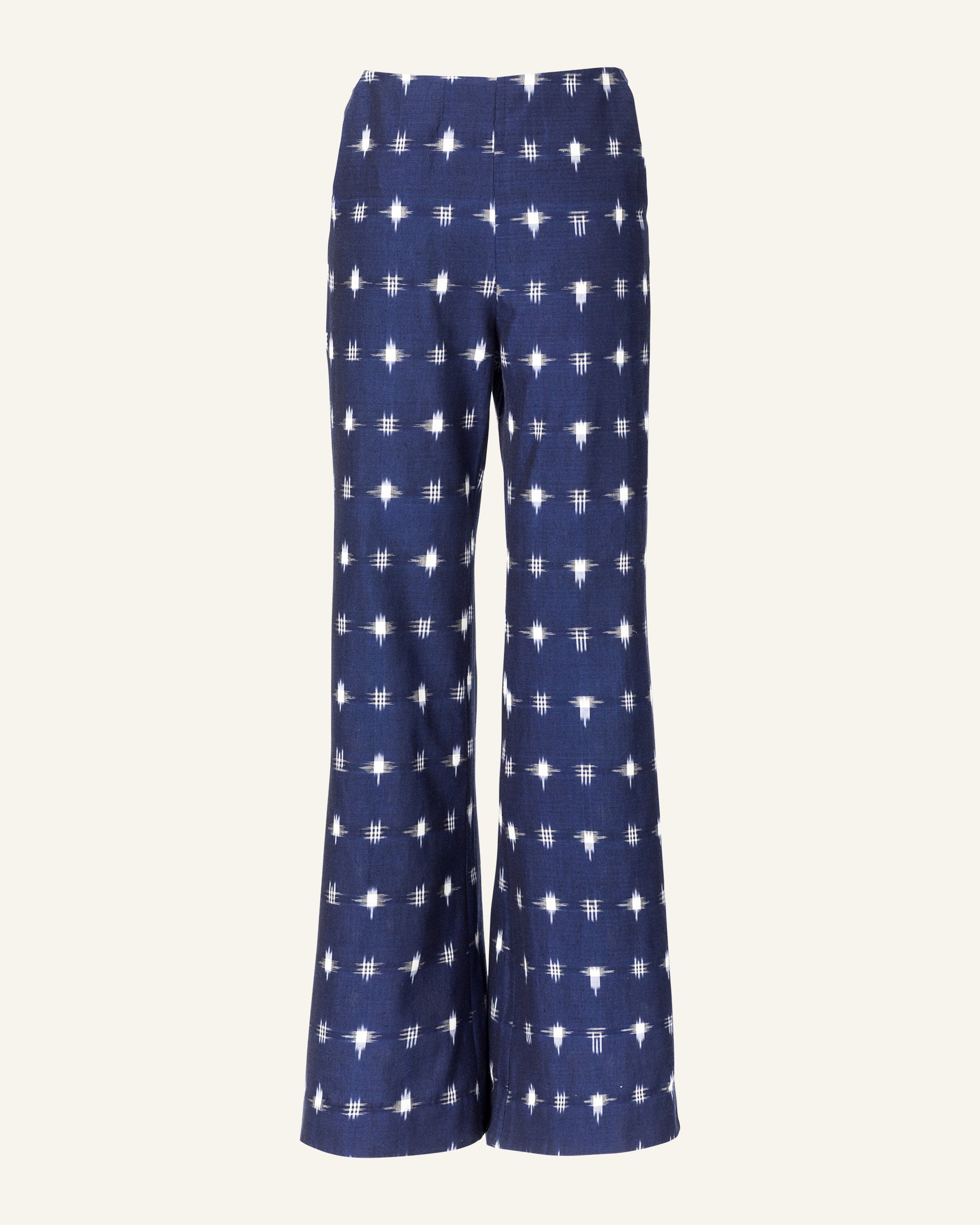 The Charles Navy Ikat Spot Pant showcases a striking white geometric pattern reminiscent of Indian ikat on navy blue fabric. These high-waisted pants with flared legs are set against a simple light backdrop, radiating classic elegance and style.