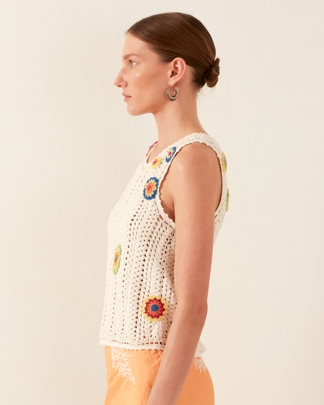 A side profile of a woman with brown hair styled in a bun, wearing the Lula Pearl Flower Crochet Top, a vintage-inspired sleeveless white tank adorned with colorful daisy motifs, paired with light orange pants. The background is a plain off-white wall.