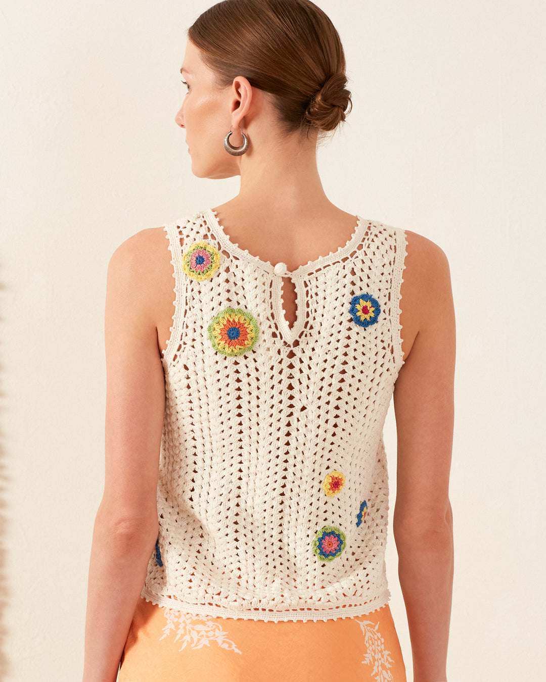 With short brown hair, a person is wearing the Lula Pearl Flower Crochet Top, a vintage-inspired piece featuring colorful circular patterns that evoke a daisy motif. The sleeveless design includes a button closure at the back as they pose against a plain white background.