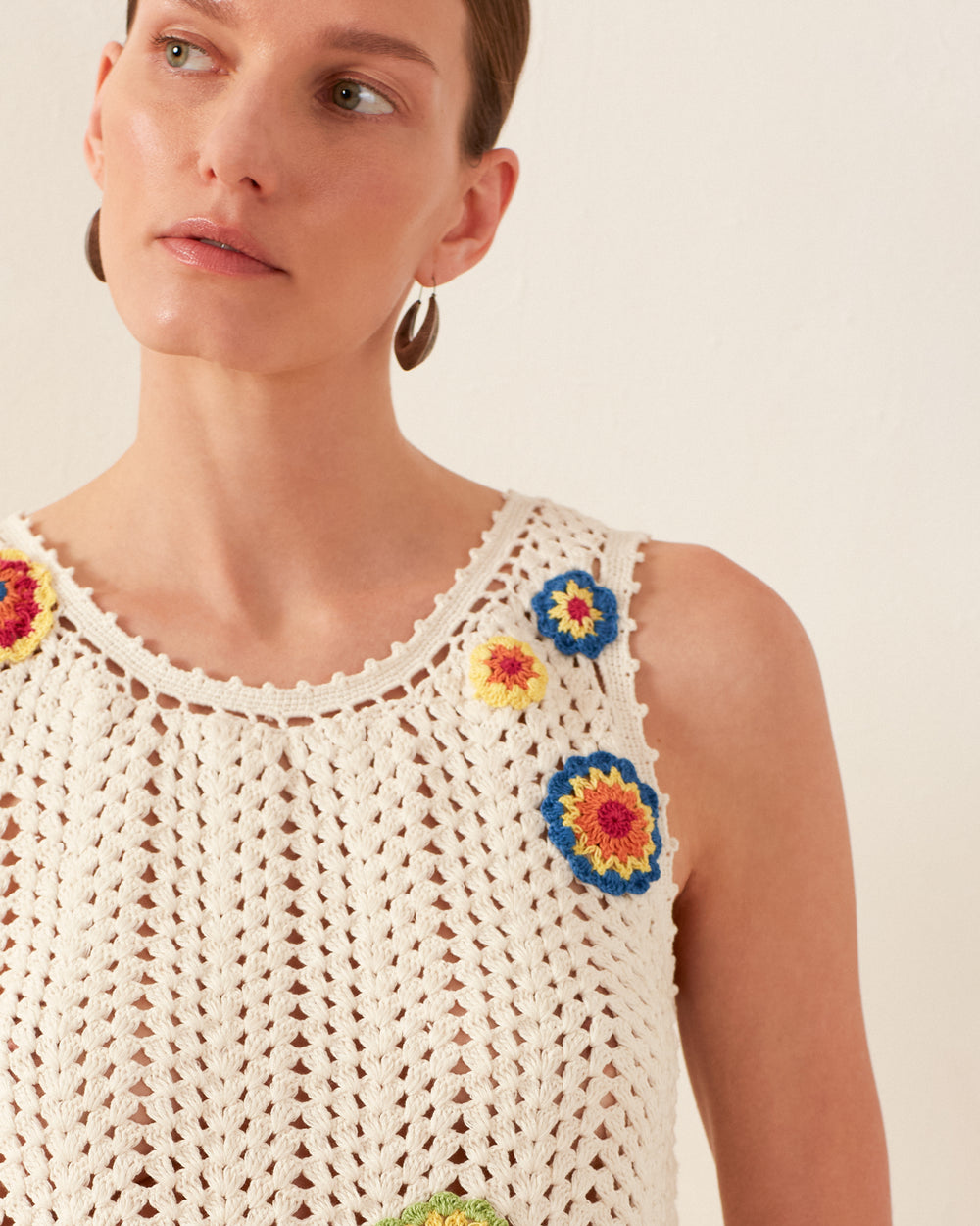 A person wearing the Lula Pearl Flower Crochet Top, a sleeveless white tank adorned with vibrant floral designs and a charming daisy motif, looks to the side. They have short hair and are wearing hoop earrings. The background is plain and light-colored, enhancing the vintage-inspired vibe.