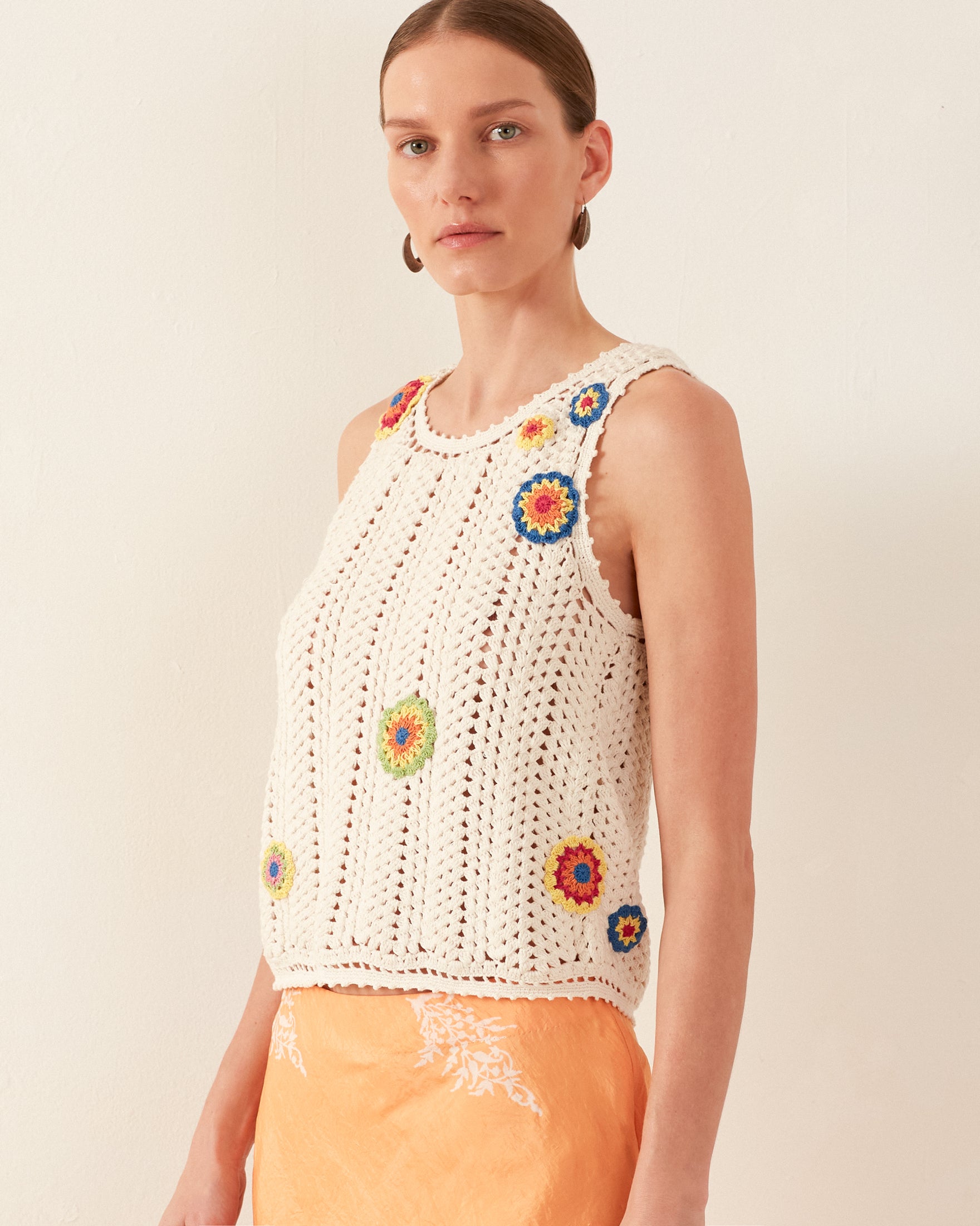 A woman models the Lula Pearl Flower Crochet Top, a vintage-inspired, sleeveless white tank intricately designed with colorful floral patterns. She pairs it with an orange skirt featuring a subtle daisy motif. Her hair is elegantly pulled back as she poses against a plain, light-colored background.