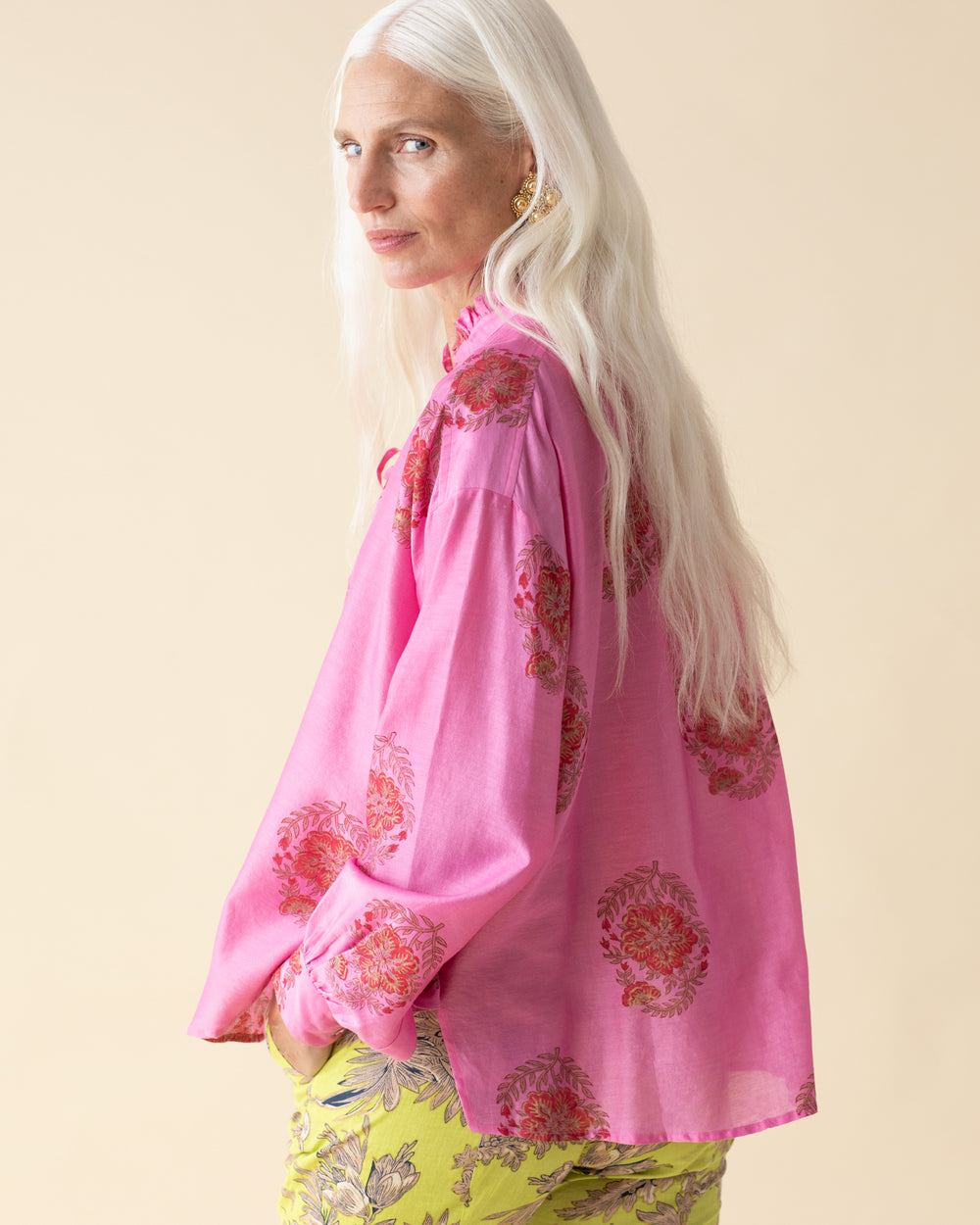 An older woman with long white hair, wearing the Willa Rosaria Silk Shirt—a pink blouse featuring a ruffled collar and floral patterns—and yellow pants, stands against a beige background, looking to her left.