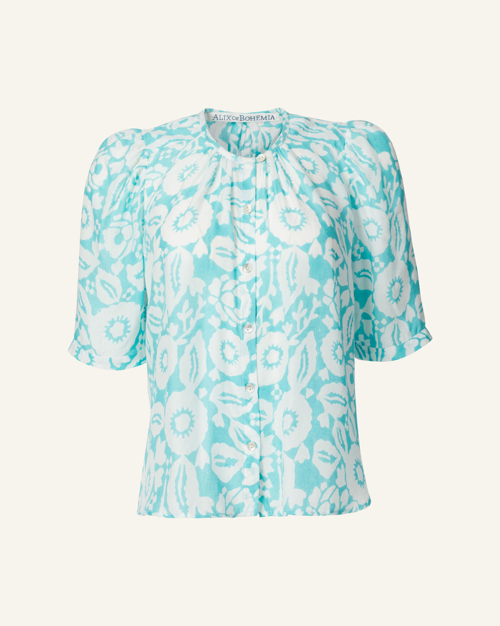 The Winona Turquoise Flower Shirt is a short-sleeved blouse featuring a white floral pattern and gathered neckline, adorned with Mother of Pearl buttons. Made from hand-printed tencel, the fabric has a light and flowy appearance, making it perfect for warm weather.