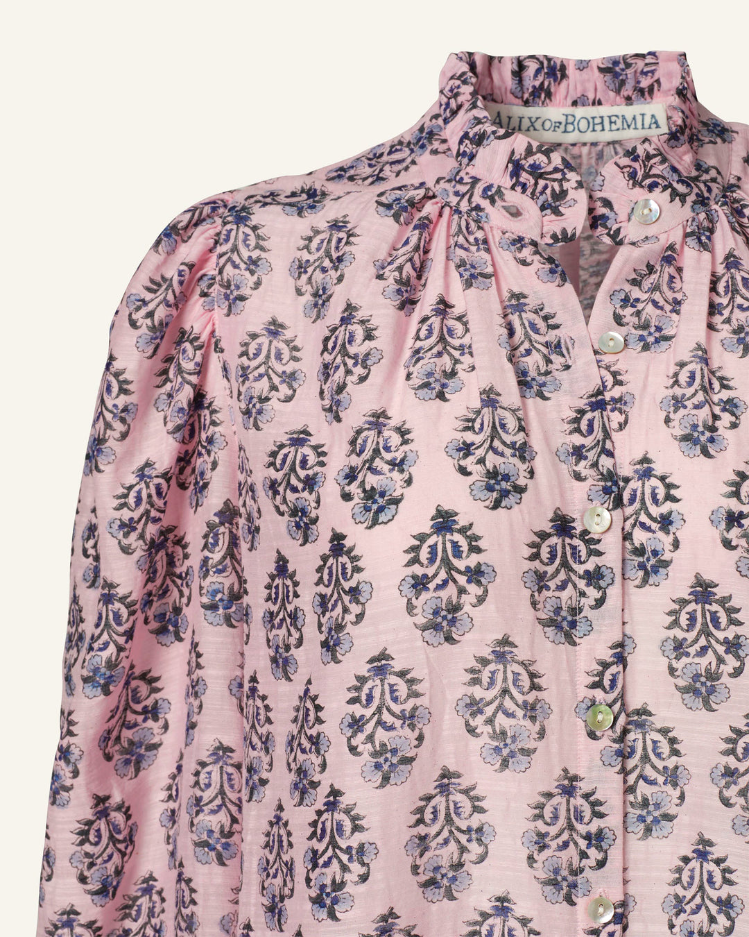 Close-up of the Annabel Pink Lily Shirt, showcasing a light pink blouse with a ruffled collar and puffed sleeves. It boasts a blue and purple floral pattern, crafted from hand-printed silk, with a button-down front. The label reads "A Piece of Bohemia.