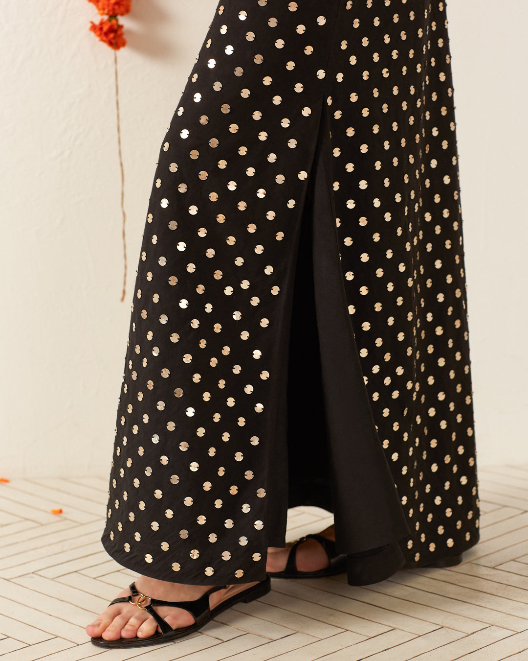 Wearing the elegant Adele Night Sky Coin Dress, featuring silver polka dot details and a side slit. Black sandals with gold accents complement the look, while a light-colored geometric design adorns the floor.